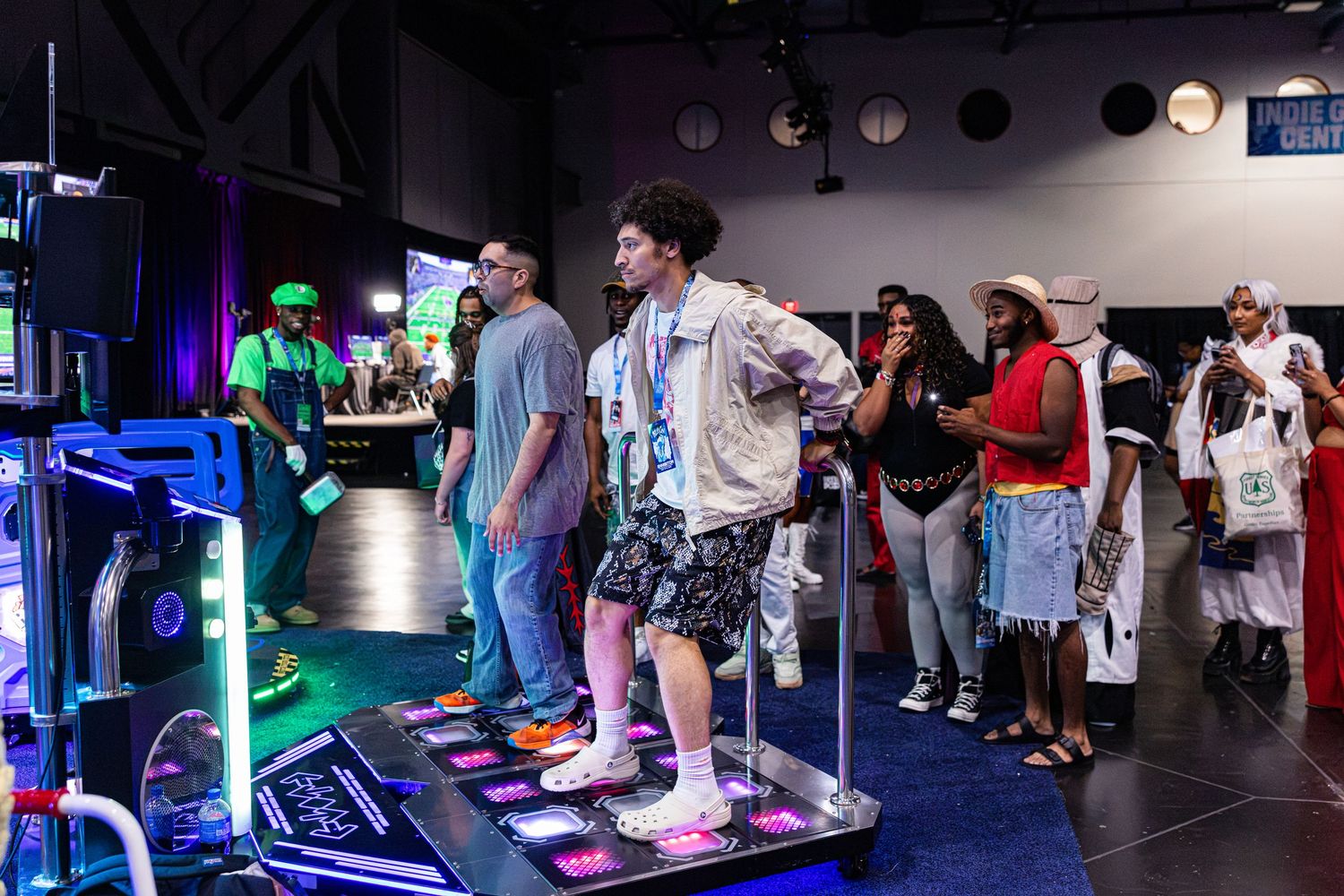 People playing Dance Dance Revolution arcade game at an indoor gaming event with spectators watching nearby.