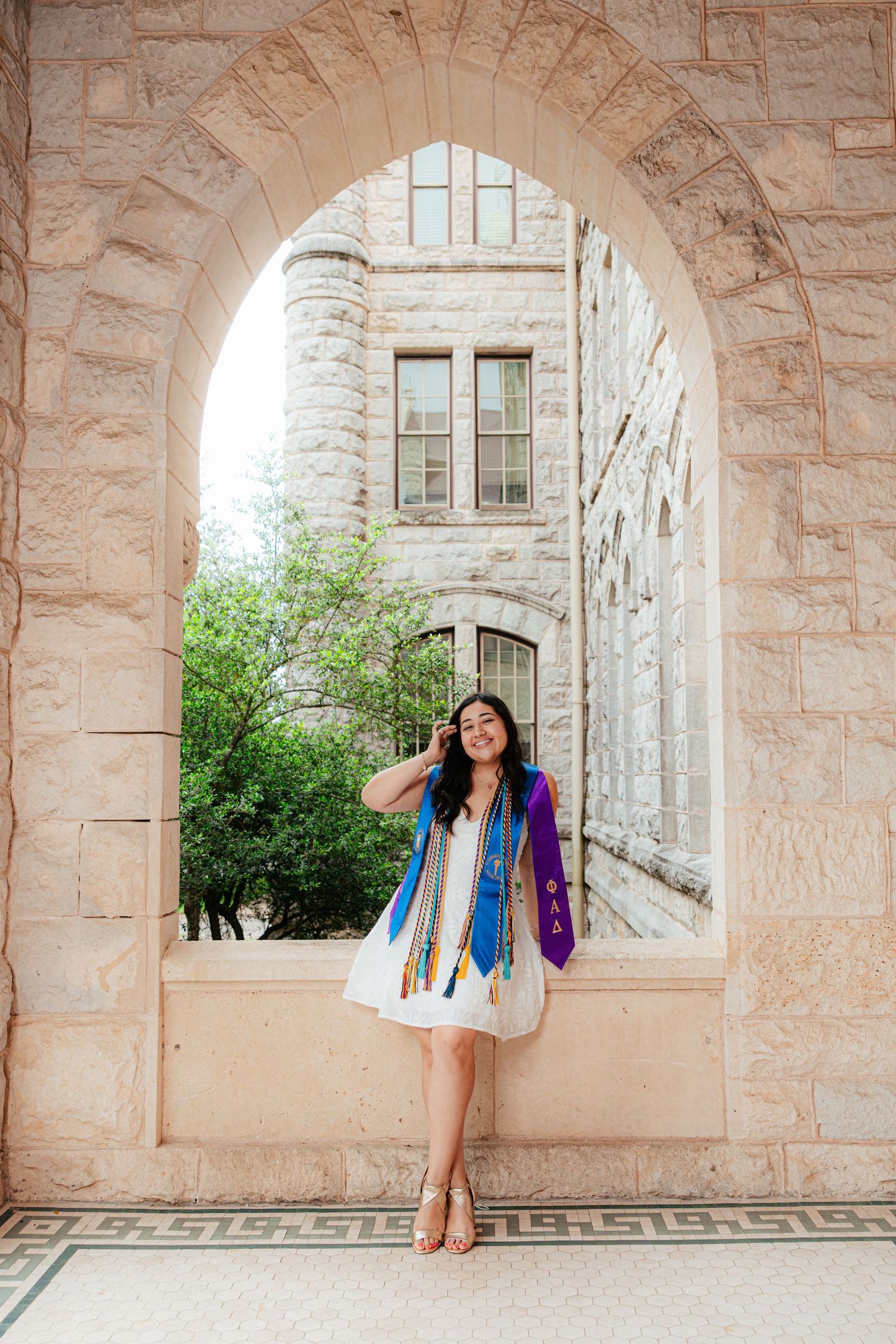 Graduate in white dress with colorful stoles poses under stone arch at university campus.