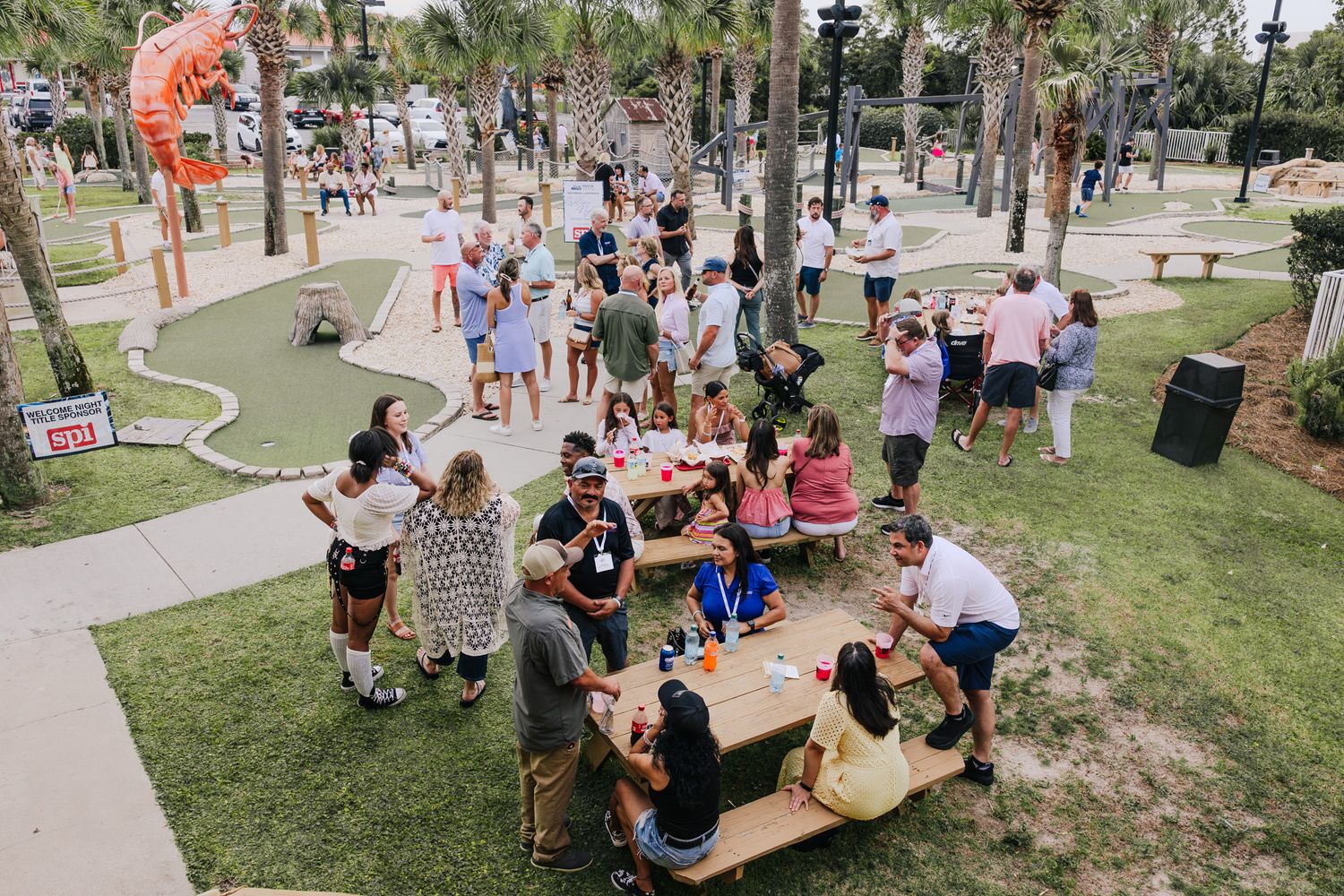 Outdoor gathering at a mini golf park with people socializing at picnic tables on a sunny day.
