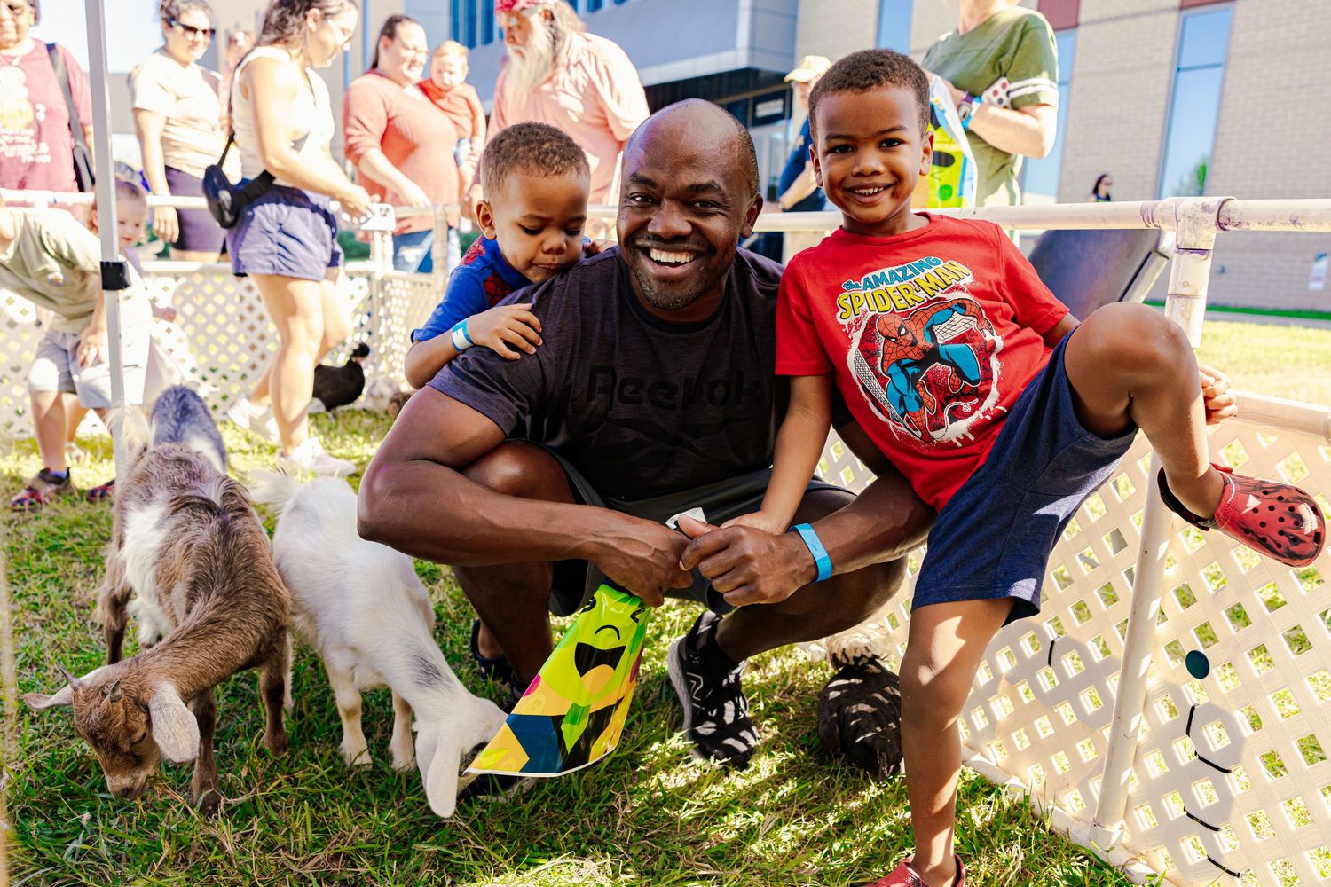 People and children pet friendly goats at an outdoor petting zoo on a sunny day.