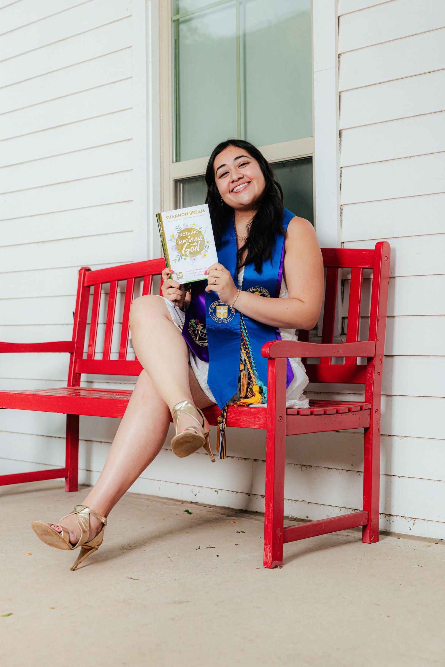 Smiling graduate in blue stole sits on red bench holding diploma, wearing gold heels, celebrating academic achievement outdoors.