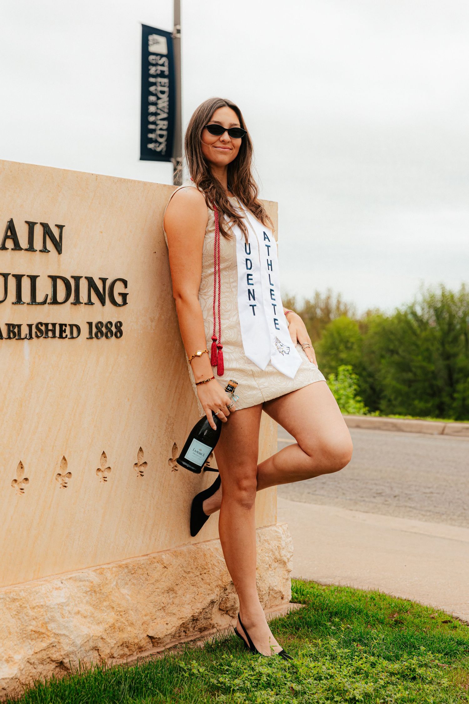 Female student athlete in graduation sash leans against a stone building sign, holding heels, wearing sunglasses outdoors.