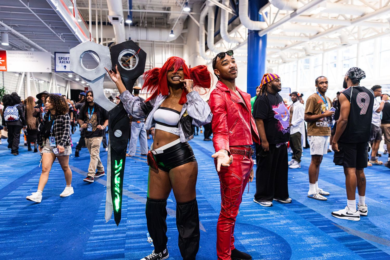 Two cosplayers in vibrant costumes pose at a comic convention on a blue carpet floor.