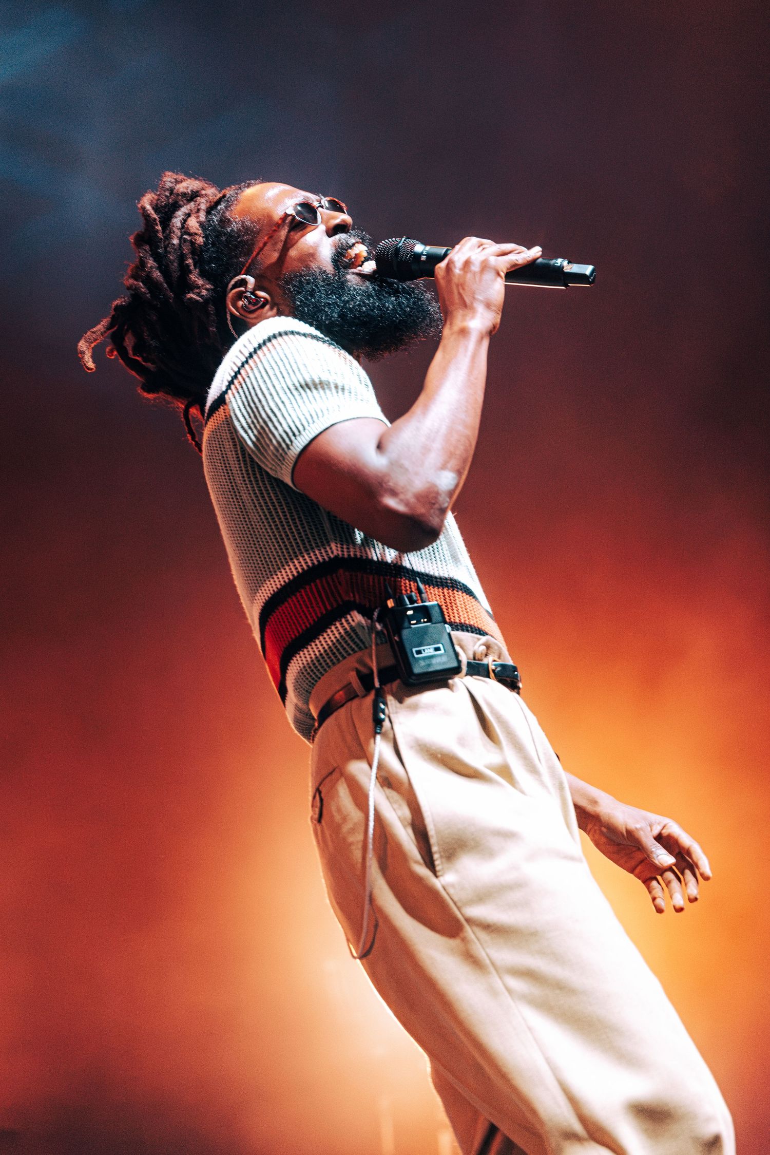Male performer with dreadlocks and beard singing passionately into microphone on stage with dramatic orange lighting.