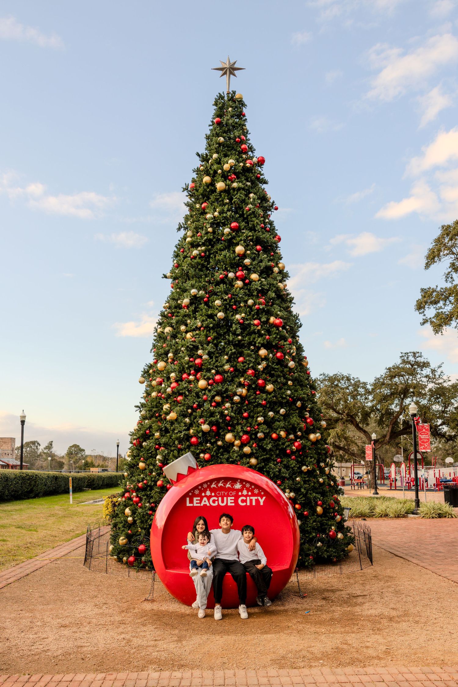 Large outdoor Christmas tree decorated with ornaments and lights next to a red League City sign display in a public plaza.
