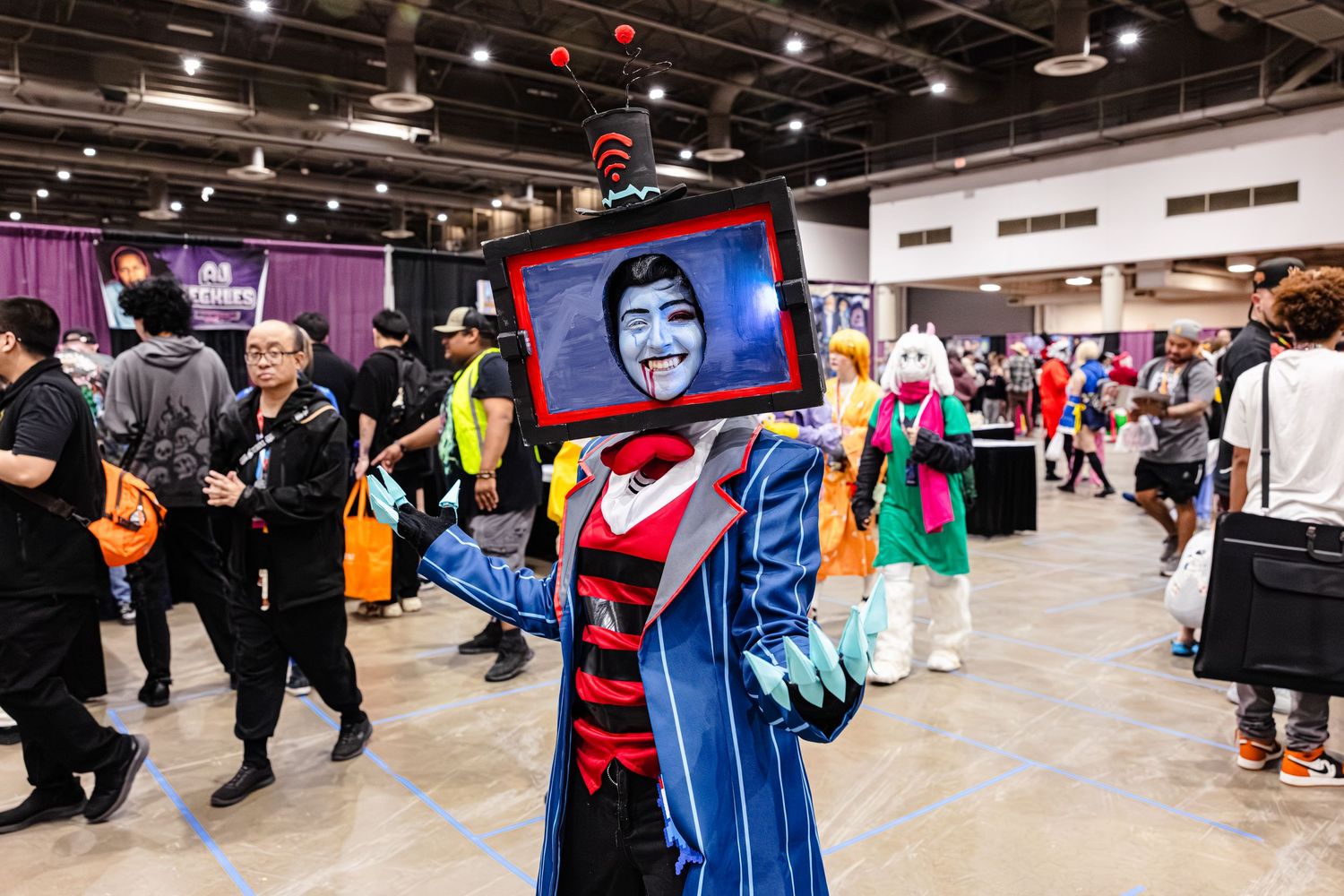 Cosplayer wearing a TV head costume at a comic convention, dressed in a blue cape with a face displayed on the screen.