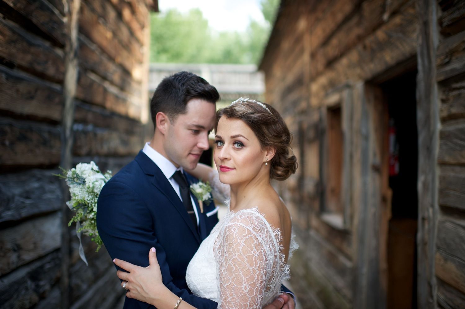 Bride and groom embracing closely between rustic wooden buildings, bride gazing toward the camera.