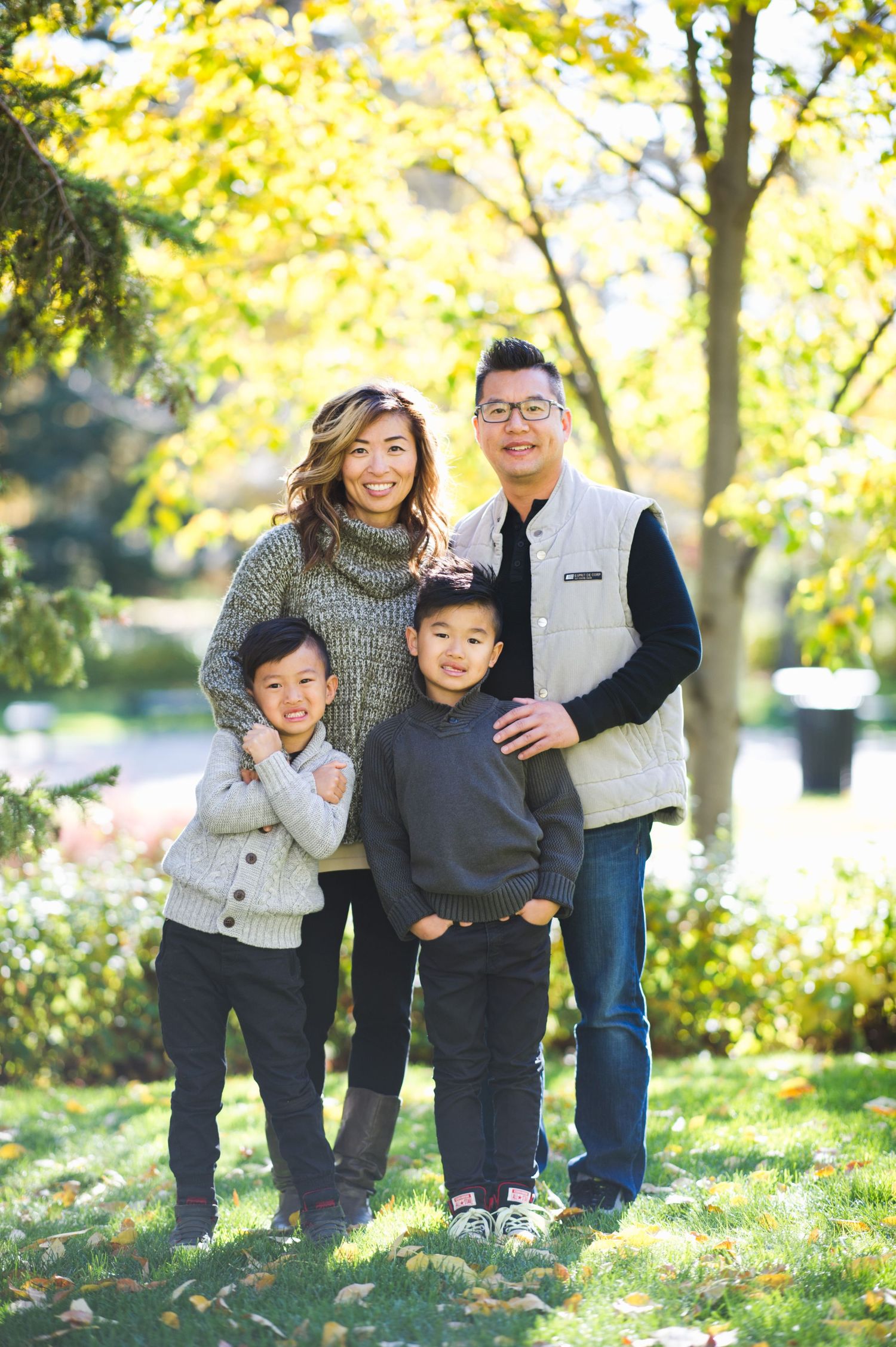 Family of four posing outdoors on a sunny day, smiling in front of bright fall trees.