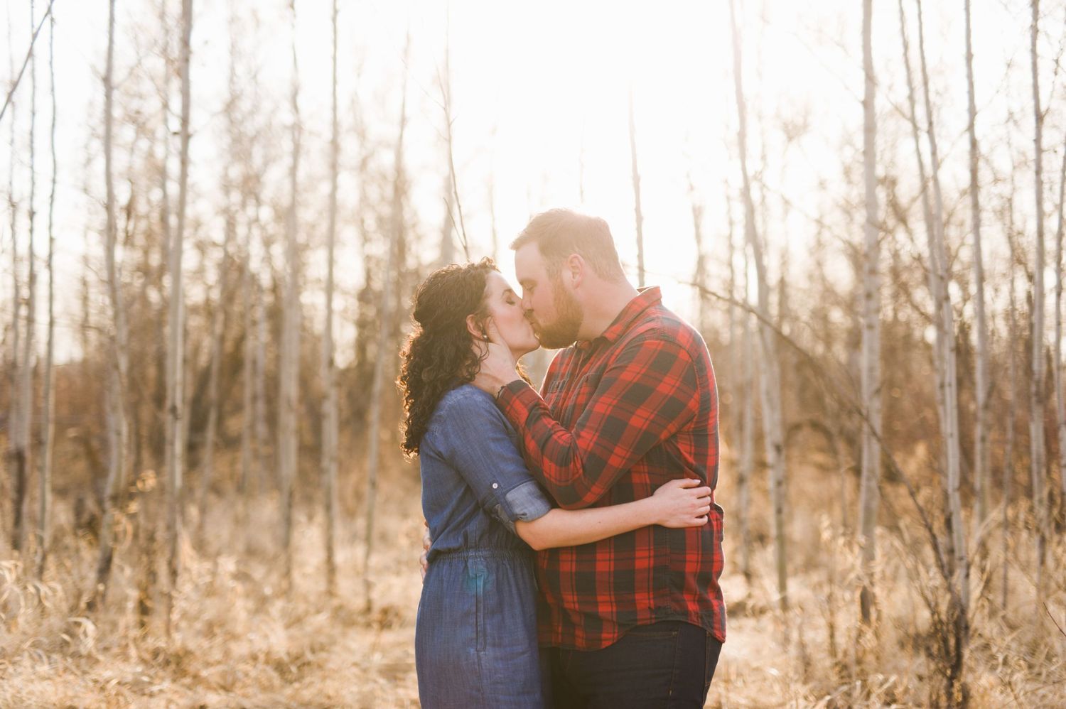 Romantic kiss shared by a couple surrounded by leafless trees in golden hour light, arms wrapped around each other in a quiet forest.