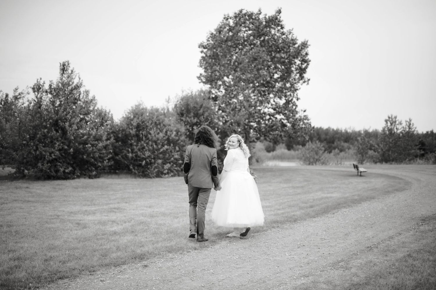 Black and white image of newlyweds walking hand in hand down a curved path surrounded by trees, captured from behind.