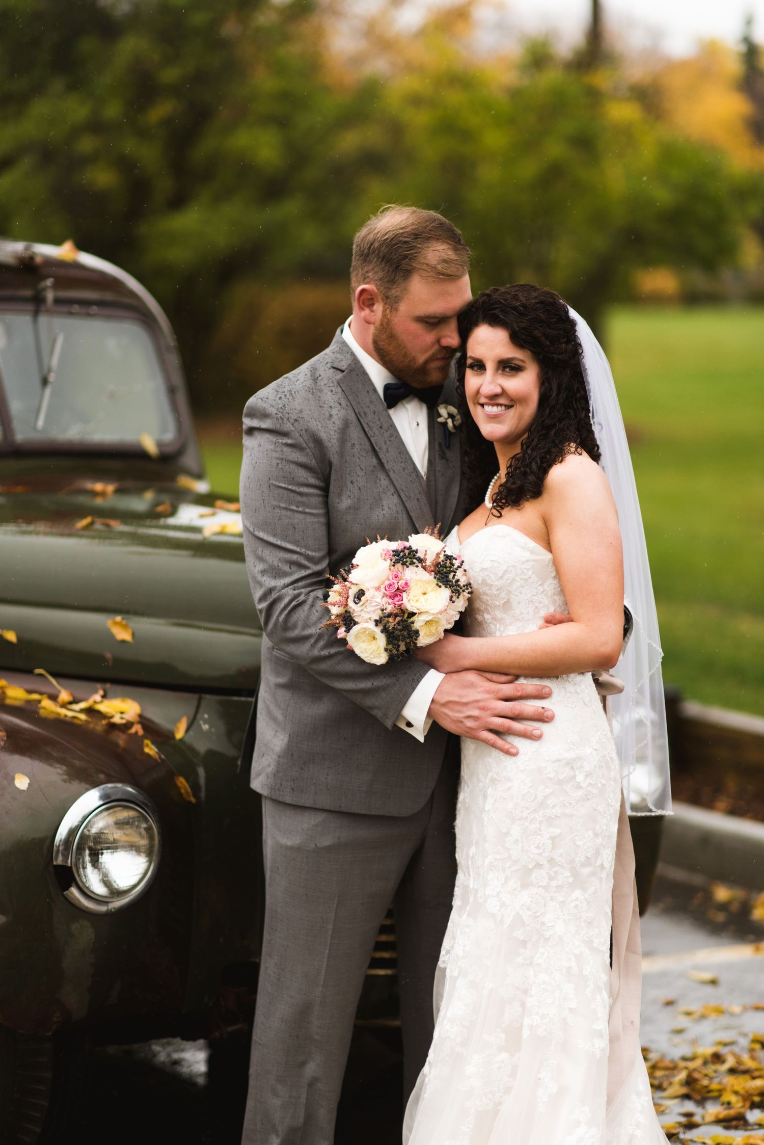 Groom wrapping his arms around his bride as she smiles gently, both standing in front of a rustic vintage pickup with fall colors behind