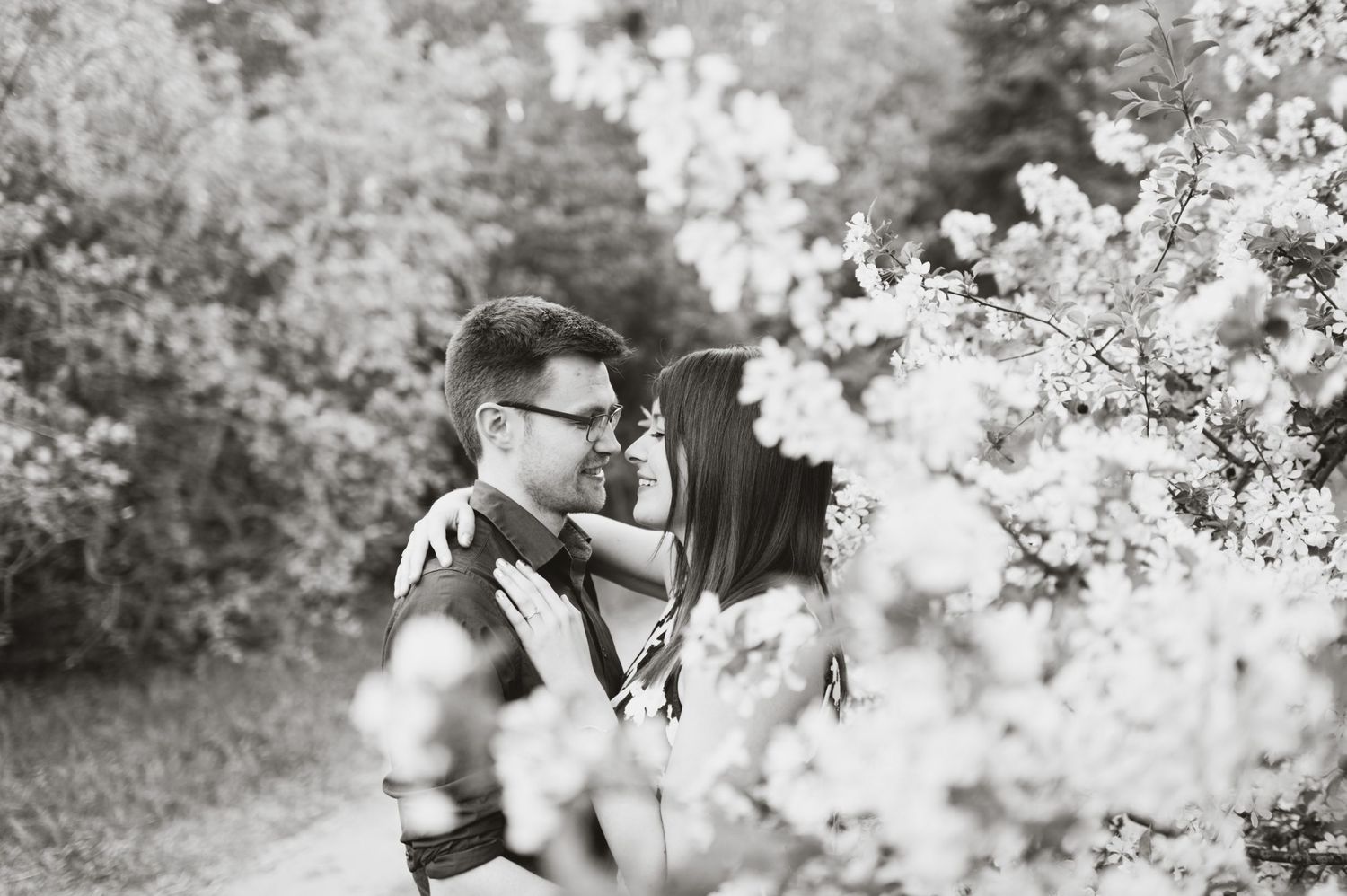 Black-and-white photo of a couple smiling and holding each other near blooming branches in a forested area.