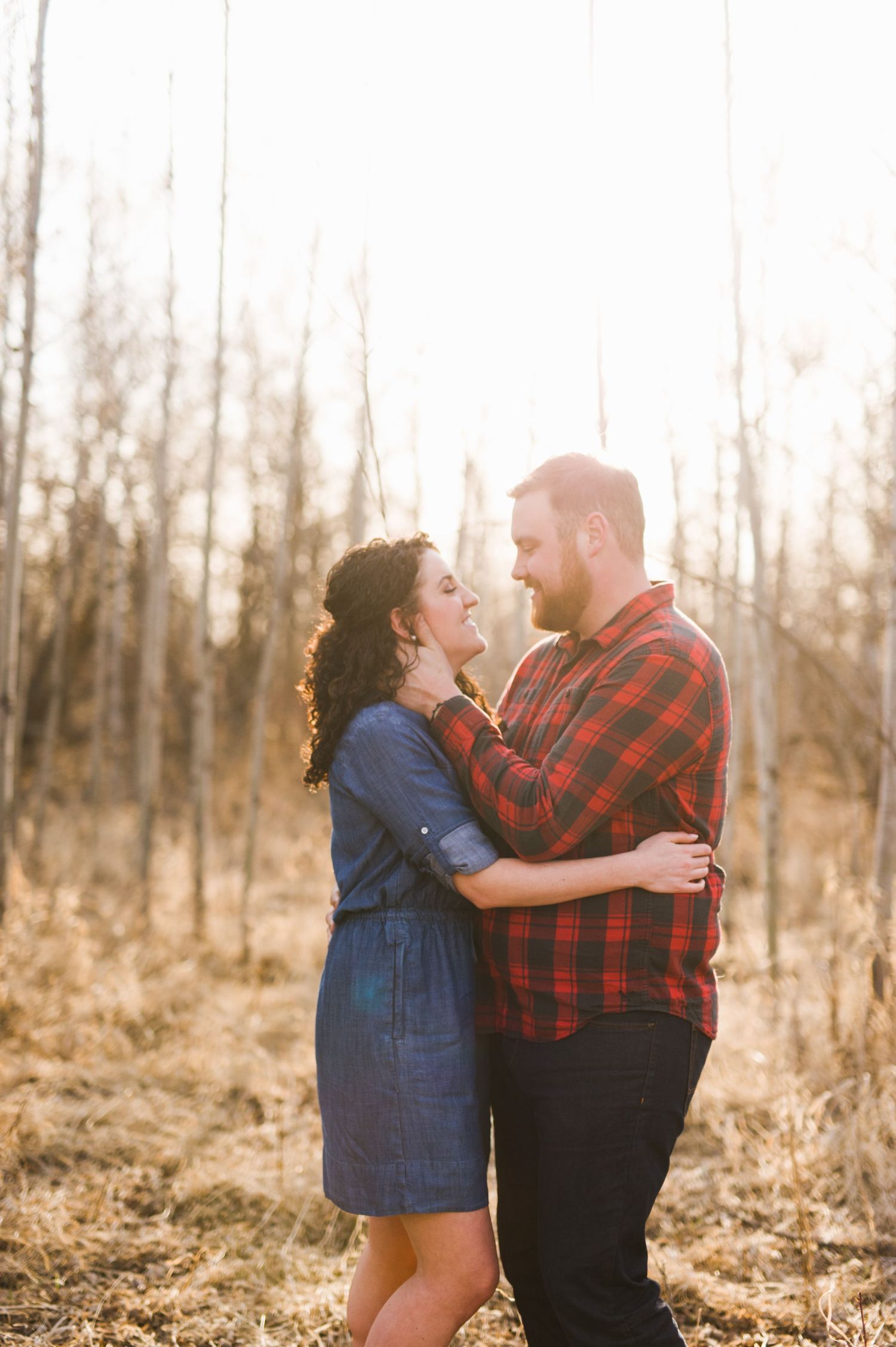 Couple holding each other in a sunlit forest, smiling and gazing into each other’s eyes — warm, golden light glowing behind them.