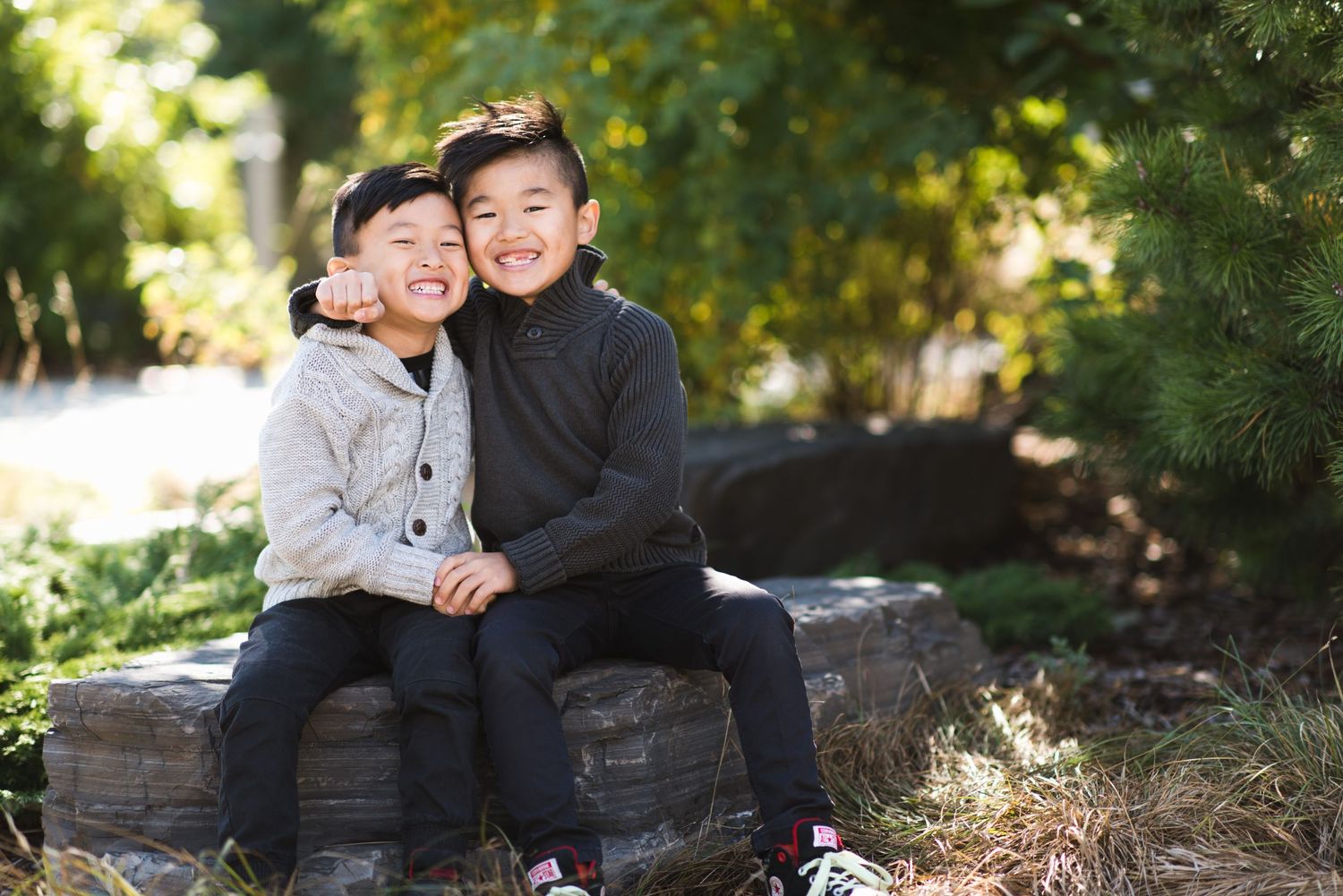 Two brothers sitting on a log, one with an arm around the other, smiling warmly at the camera.