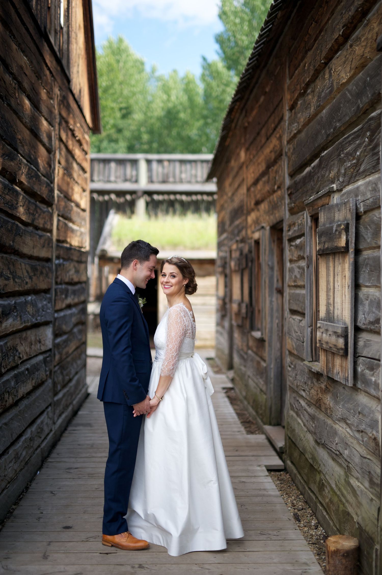 Couple stands forehead to forehead in a narrow wooden alley, holding hands and smiling softly at each other.