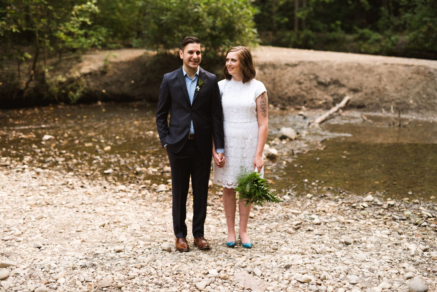 Eloping couple stands hand in hand on a rocky creek bed. Outdoor micro-wedding near river in Alberta forest.