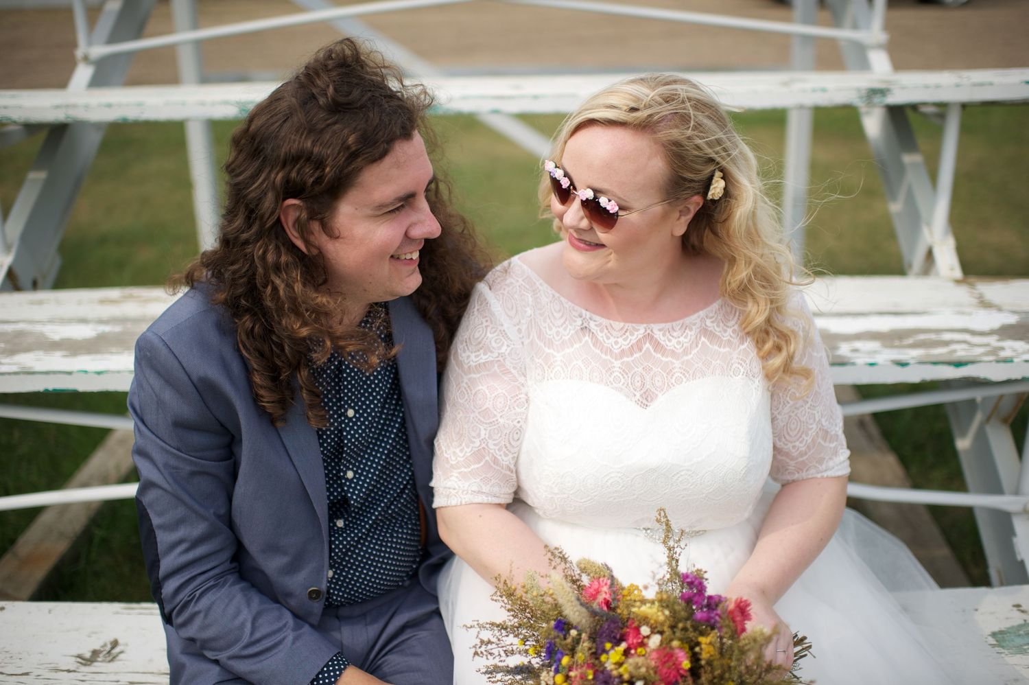 Bride wearing sunglasses and holding a wildflower bouquet, sitting with her partner on rustic bleachers, both smiling warmly.