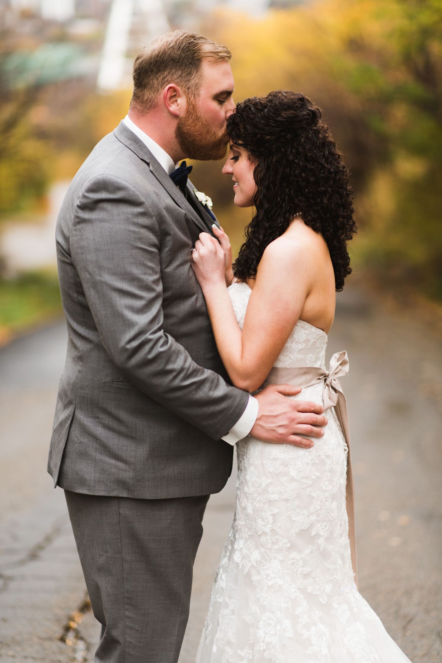 Bride and groom sharing a soft moment under a city overpass, holding each other closely along a damp path surrounded by golden trees.