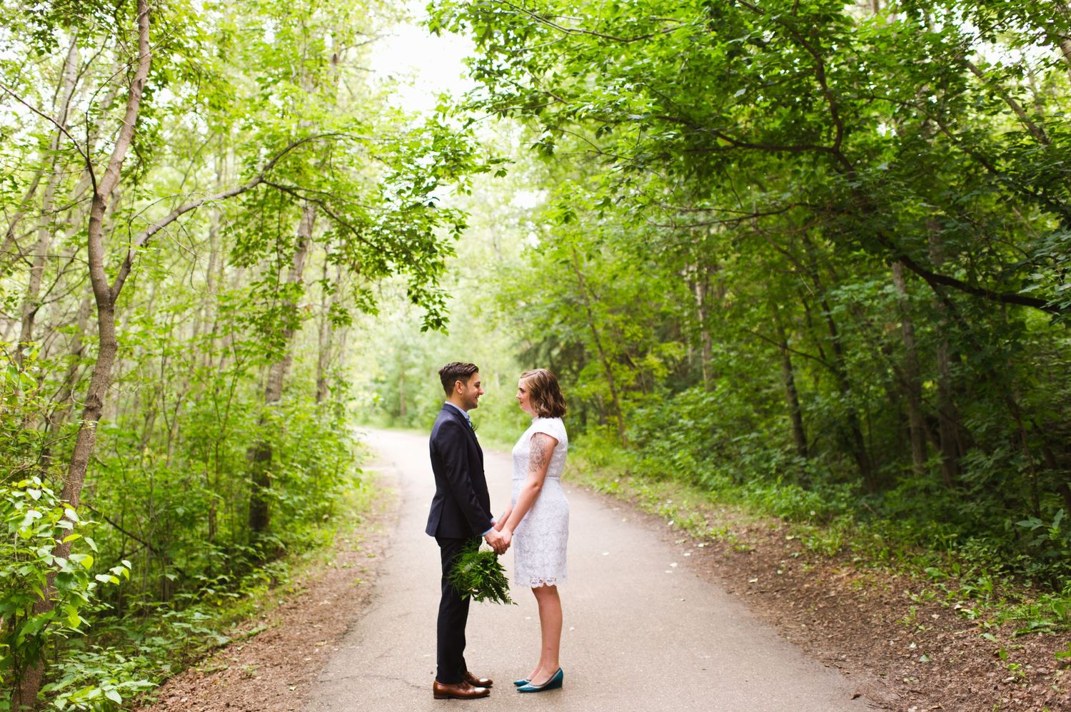 Couple stands face to face on a tree-lined path. Intimate Alberta elopement in lush, sunlit forest setting.