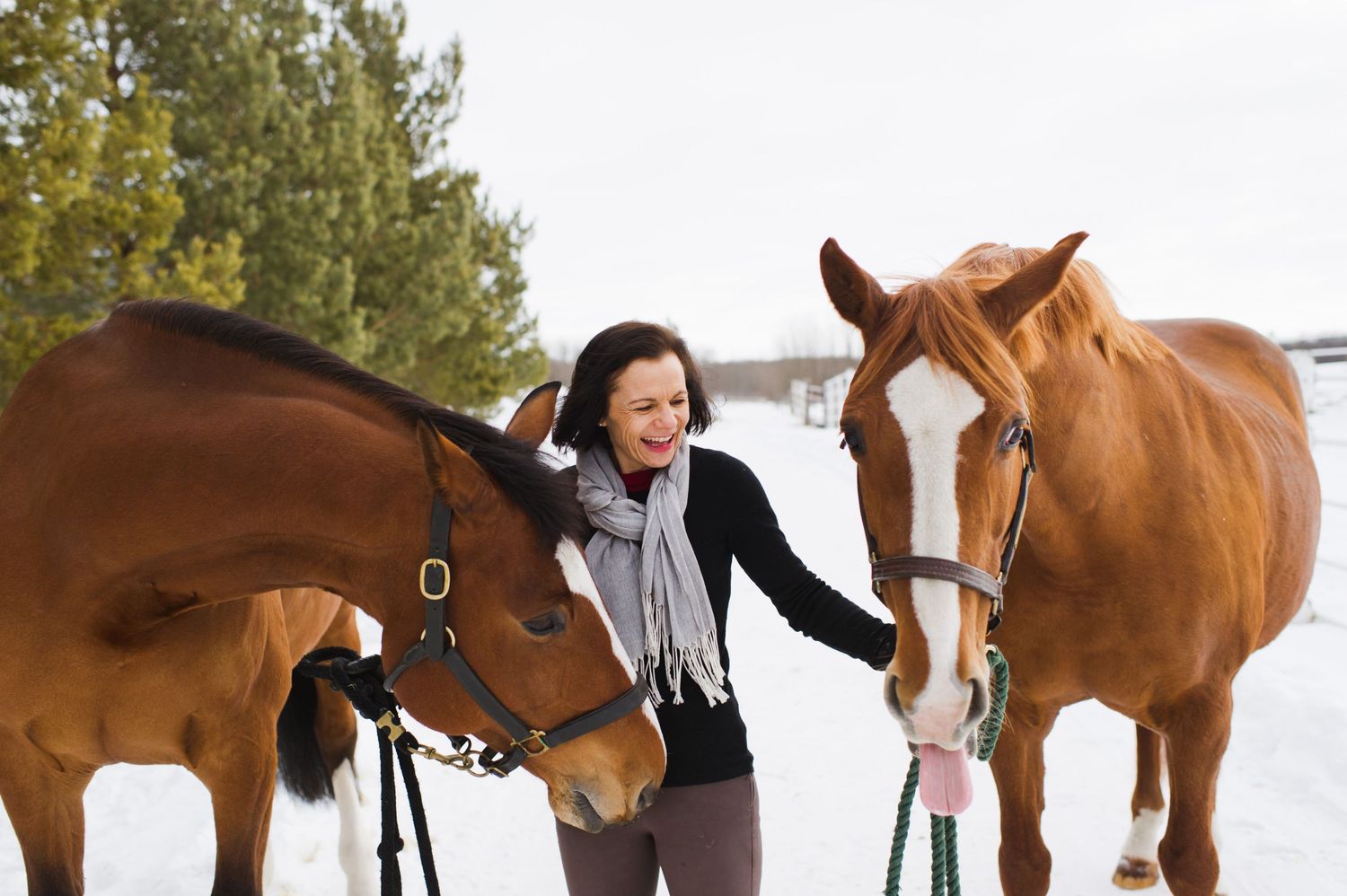 Woman standing in the snow holding two horses by their reins, laughing between them.