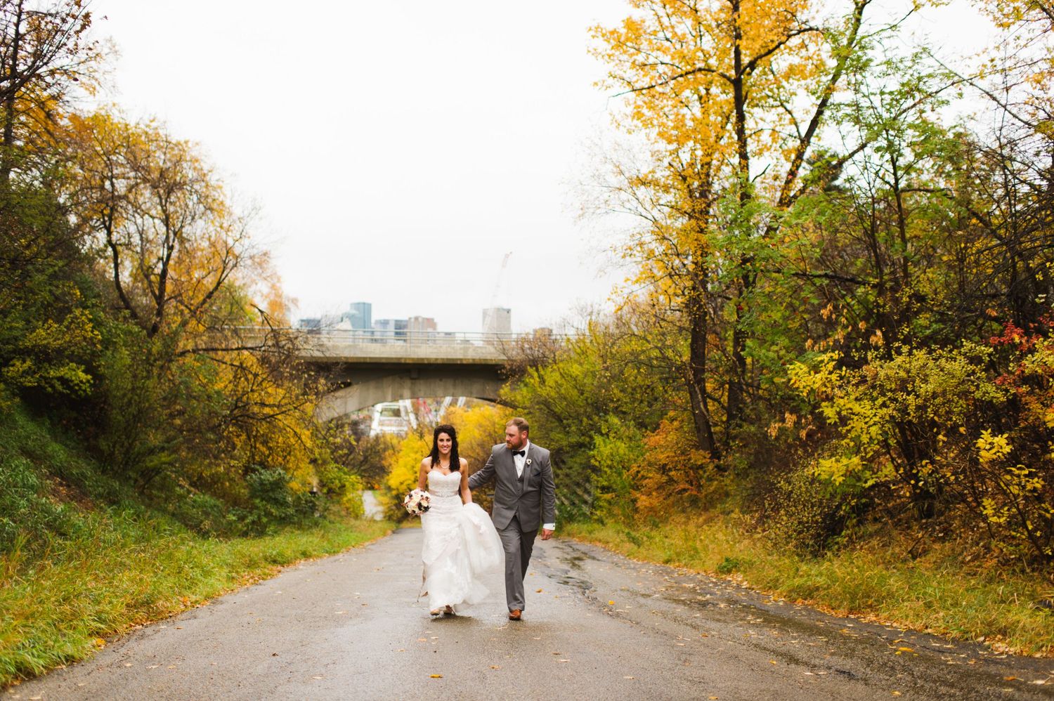 Wedding couple walking hand-in-hand down a damp path surrounded by autumn trees, city skyline visible in the distance.
