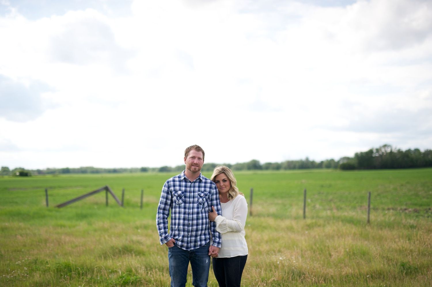 Couple standing together in an open prairie field under a wide sky, smiling at the camera. Alberta engagement photo with rustic charm.