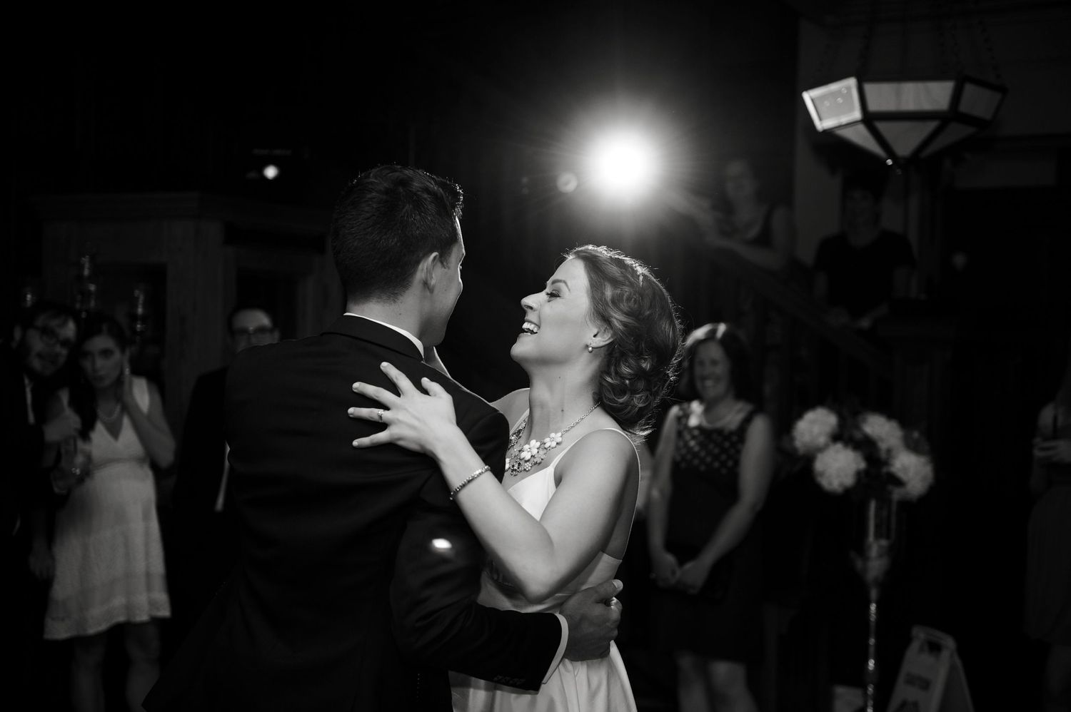 Black-and-white photo of bride smiling up at groom during their first dance, surrounded by guests.