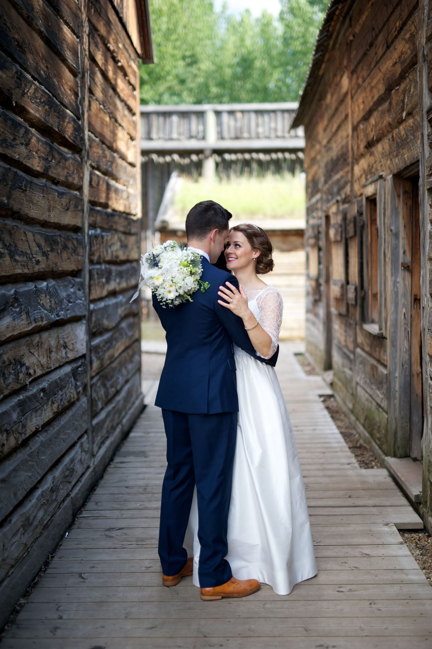 Bride and groom locked in an affectionate hug, bouquet in hand, framed between weathered wood buildings.