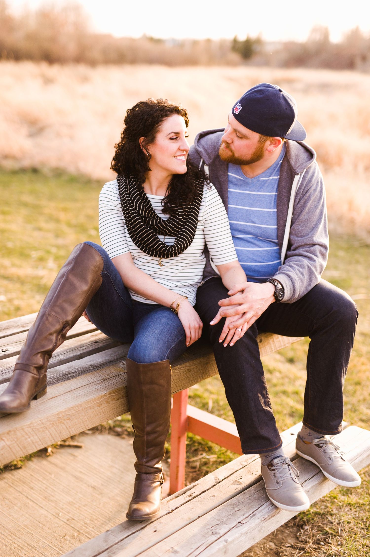 Couple sitting close together on a wooden bench in a grassy field at golden hour, gazing at each other and smiling in soft evening light.