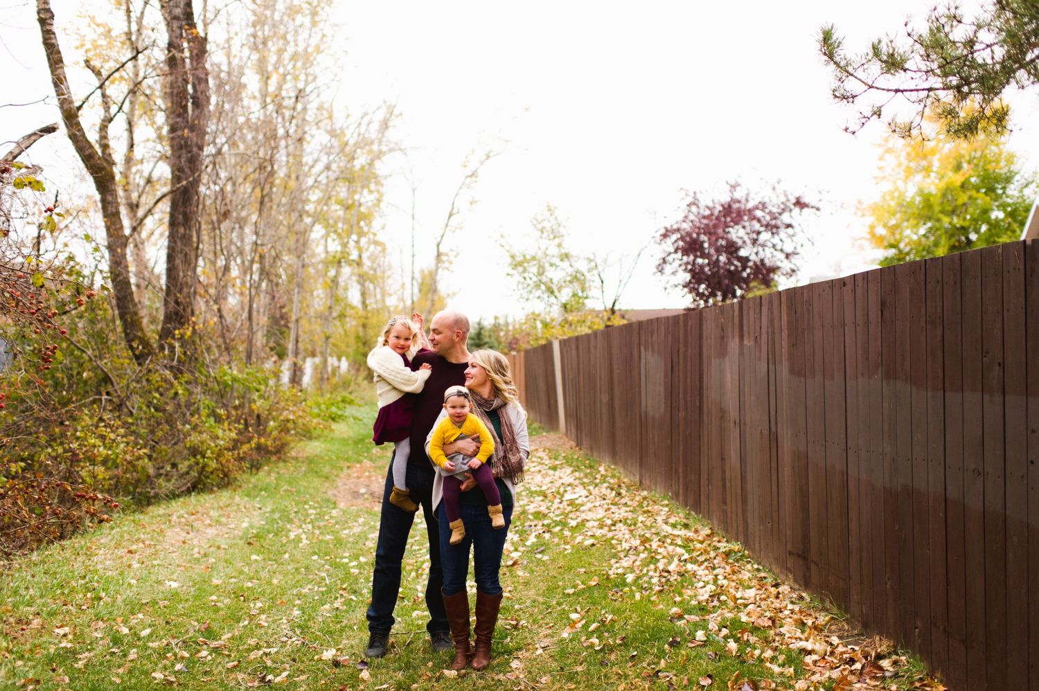 Young family walking down a leaf-covered path, carrying children through an autumn lane beside a wooden fence and colorful trees.