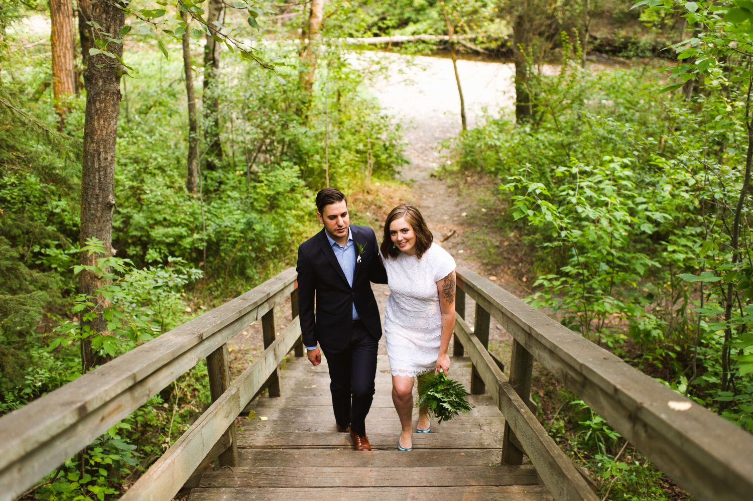 Couple walks up forest boardwalk holding hands after elopement. Nature-based Calgary micro-wedding photography.