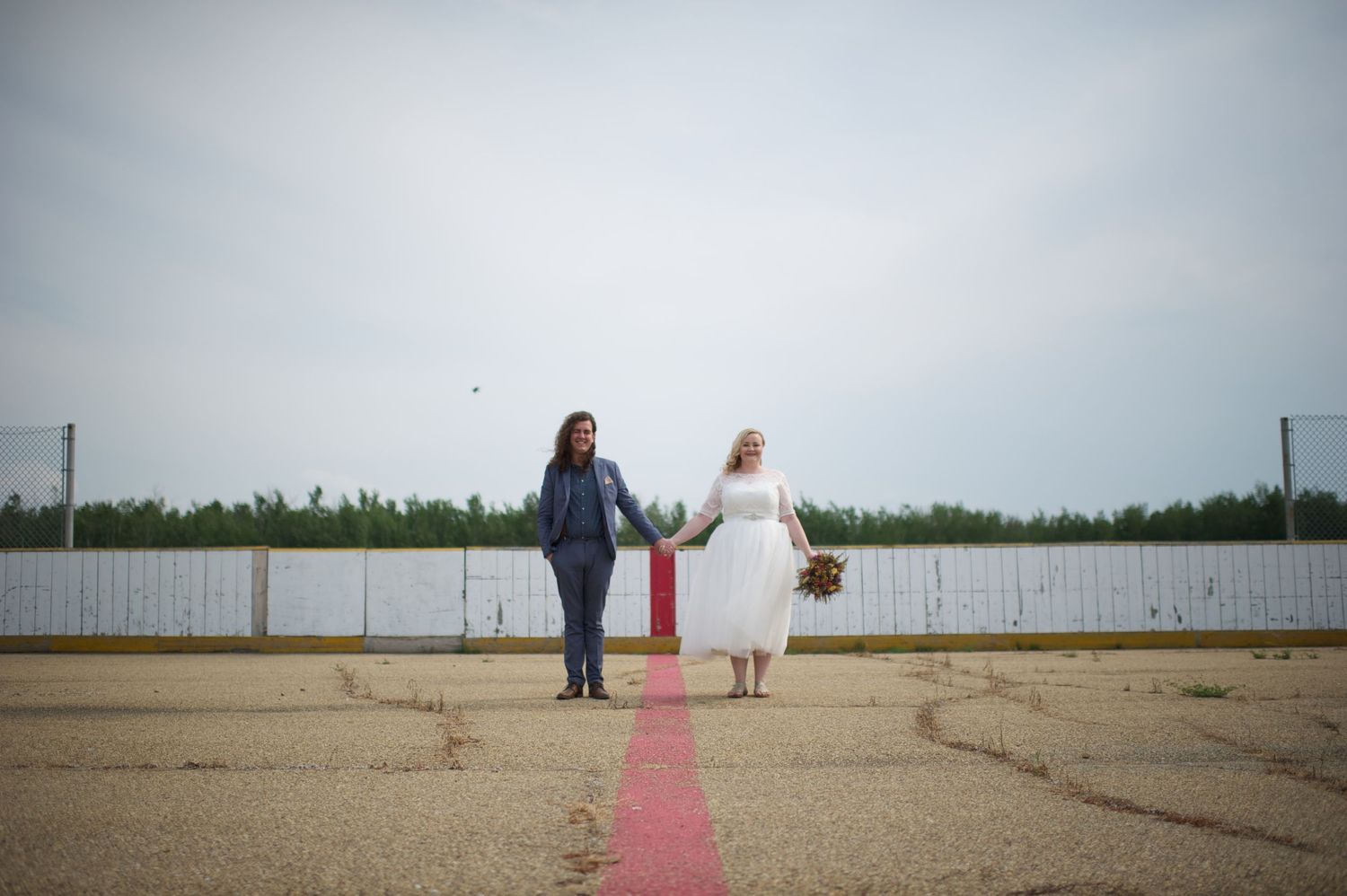 Newlyweds walking hand in hand along the center red line of an outdoor hockey rink, bride holding bouquet in one hand.