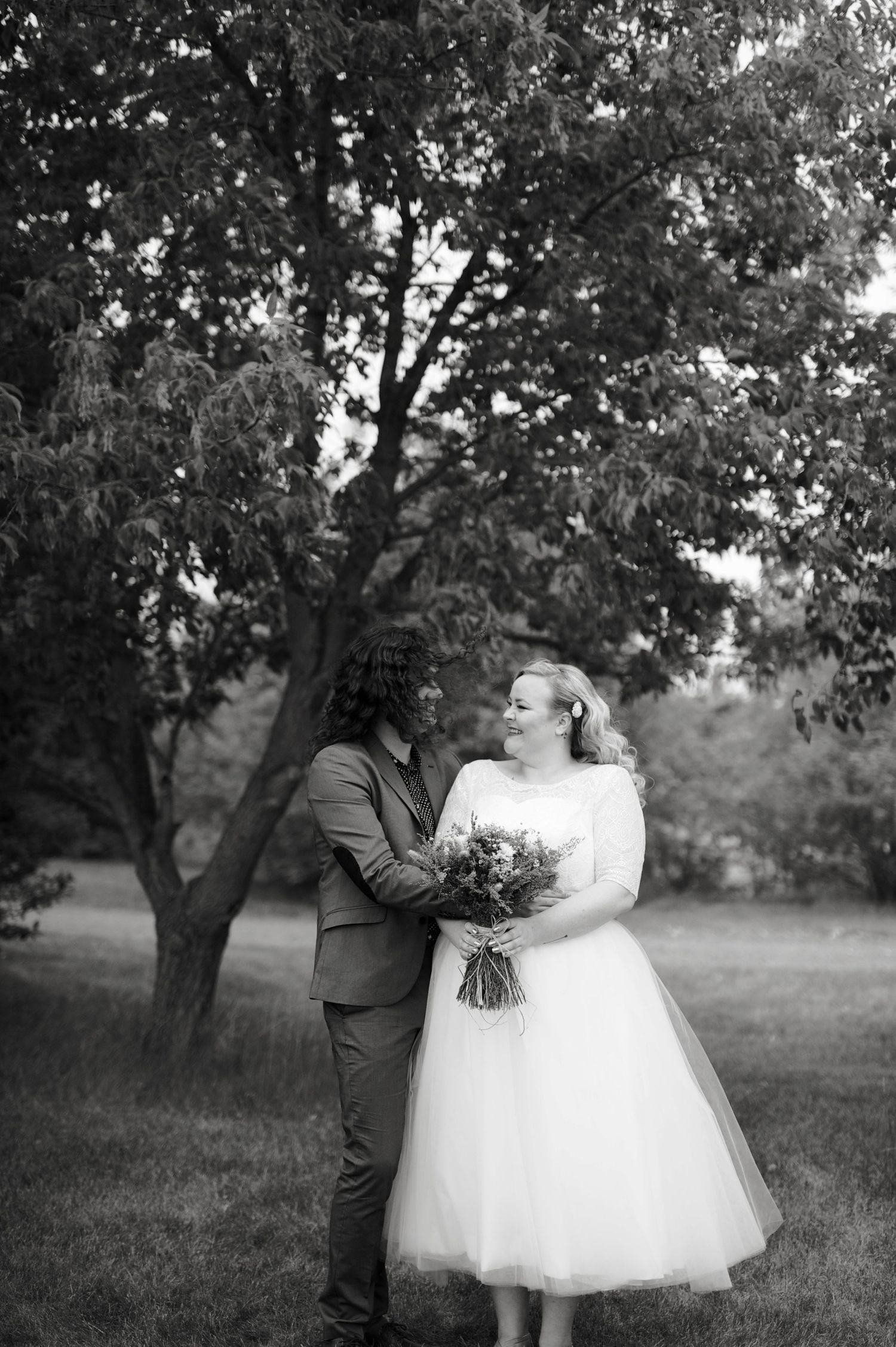 Vertical black and white wedding portrait of couple standing in a grassy park, surrounded by leafy trees.