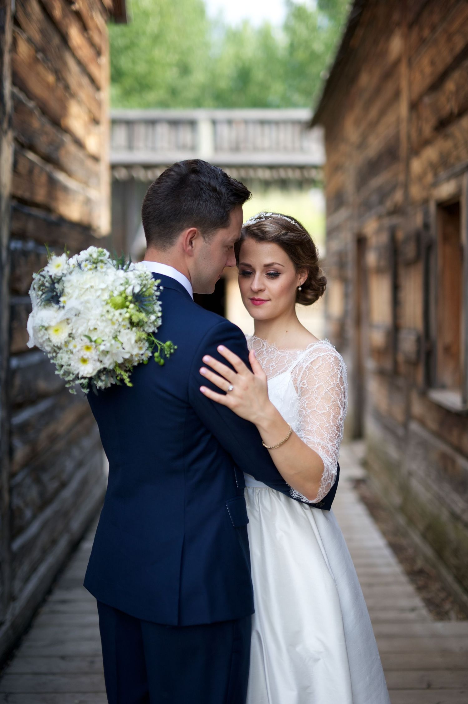 Groom holds bouquet over bride’s shoulder as she closes her eyes, resting her head against his in a rustic wooden alley.