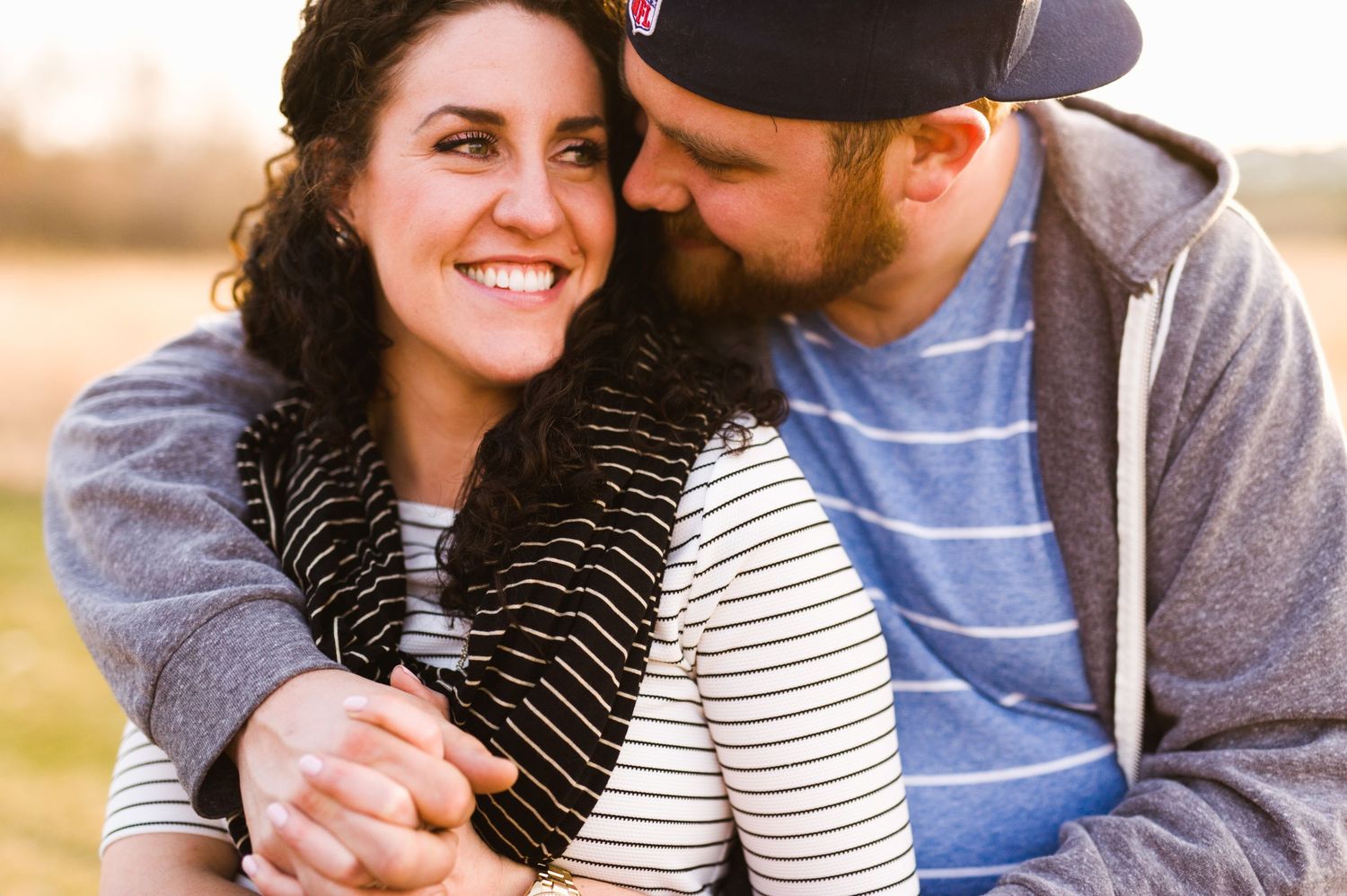 Warm sunset photo of a couple cuddling in a field, woman smiling while man kisses her cheek, dressed casually in fall layers.