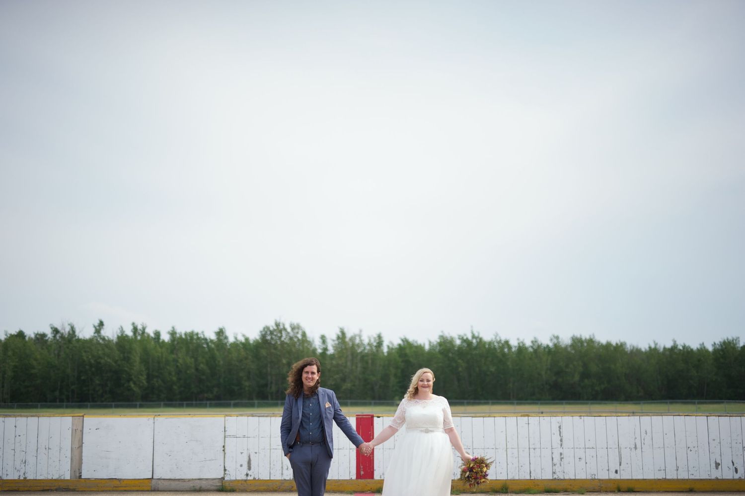 Bride in a tea-length dress and groom in a blue suit holding hands and walking across an outdoor hockey rink, bouquet in hand.