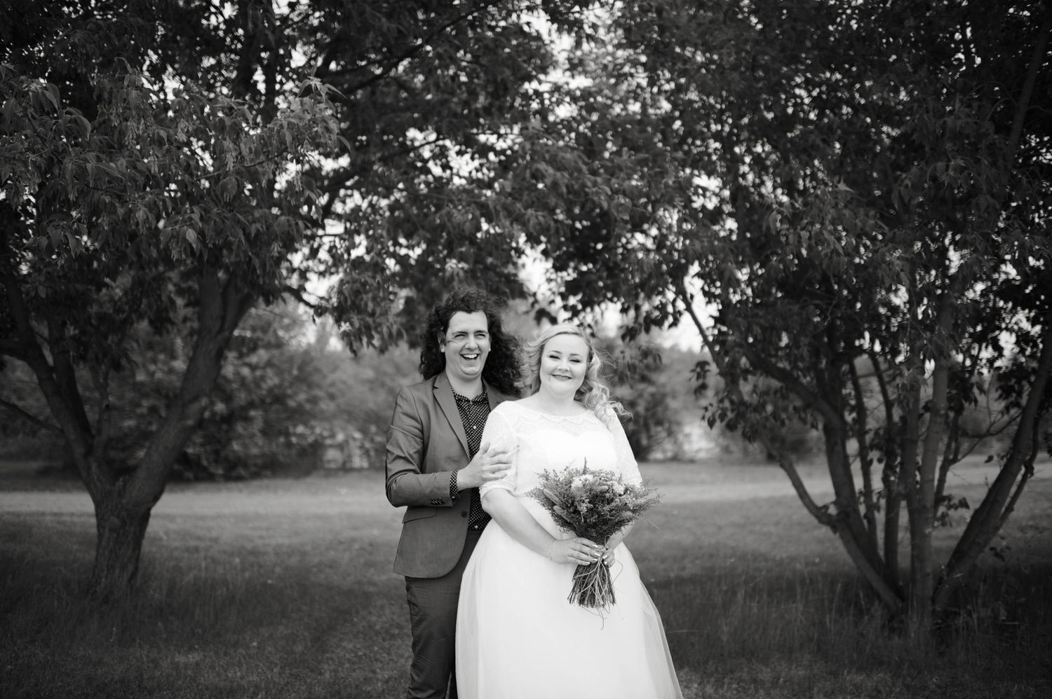 Newlyweds posed together in a quiet grove, bride holding bouquet with groom’s arm around her in a soft black and white image.