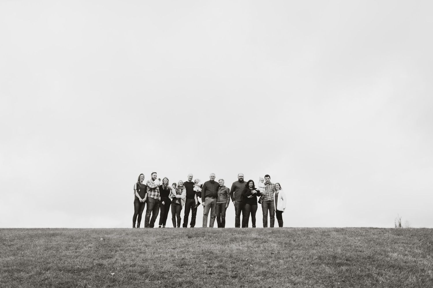 Black and white photo of an extended family standing in a line atop a grassy hill under an overcast sky, all smiling toward the camera.