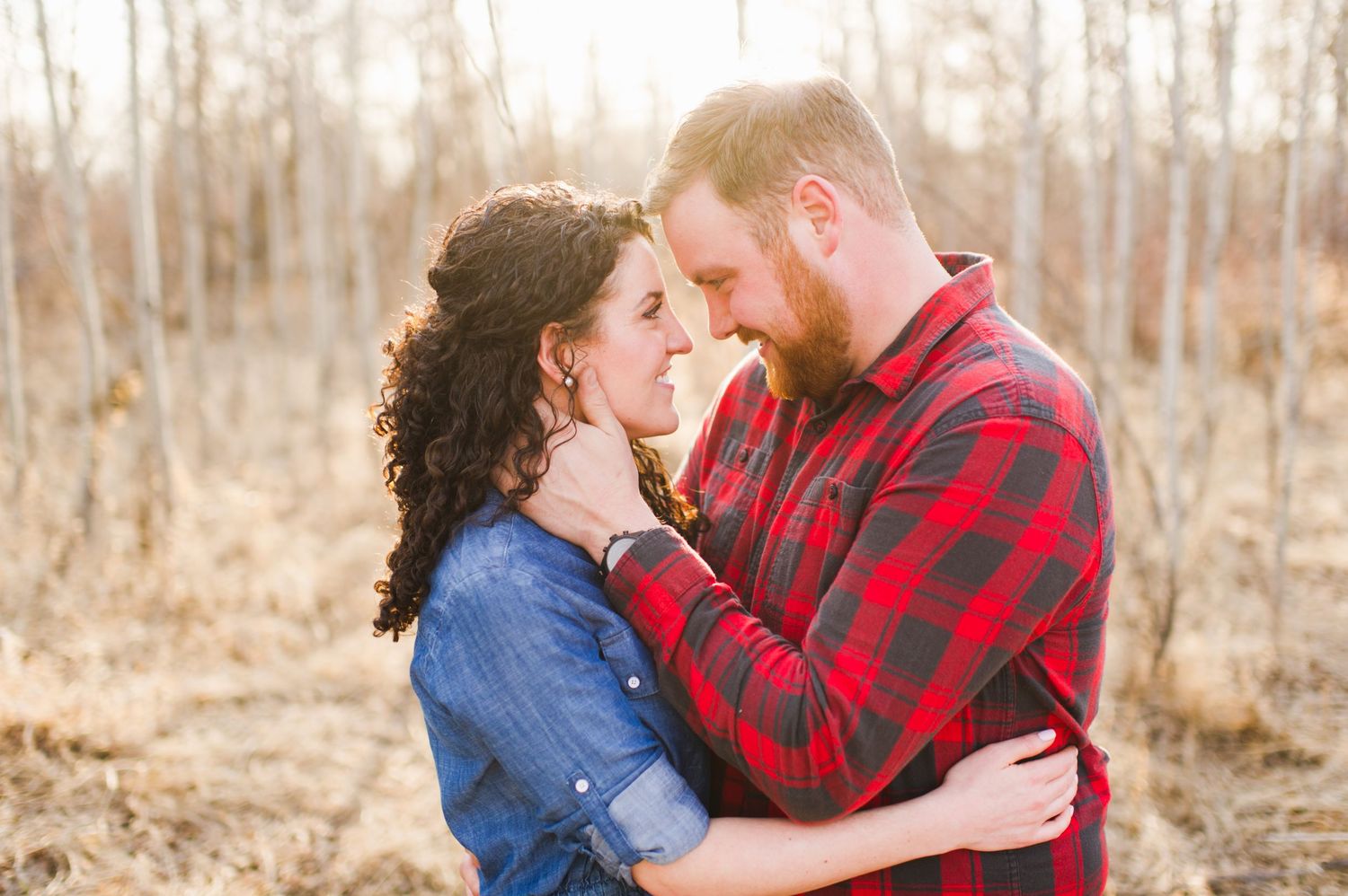 Couple standing forehead to forehead in a leafless forest, wrapped in a quiet, intimate moment with golden sunlight filtering through.