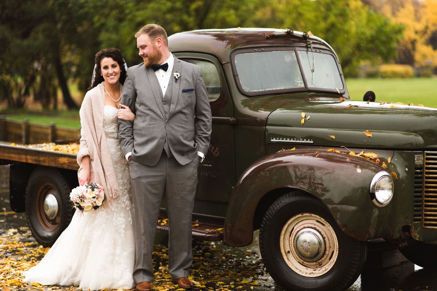 Bride holding her bouquet and shawl as she walks hand-in-hand with the groom beside a dark green vintage truck parked on a bed of leaves
