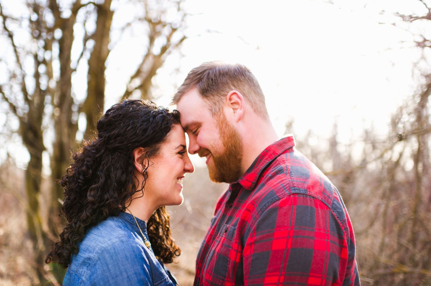 Couple sharing an intimate forehead touch, smiling and relaxed under bare trees with light filtering through behind them.