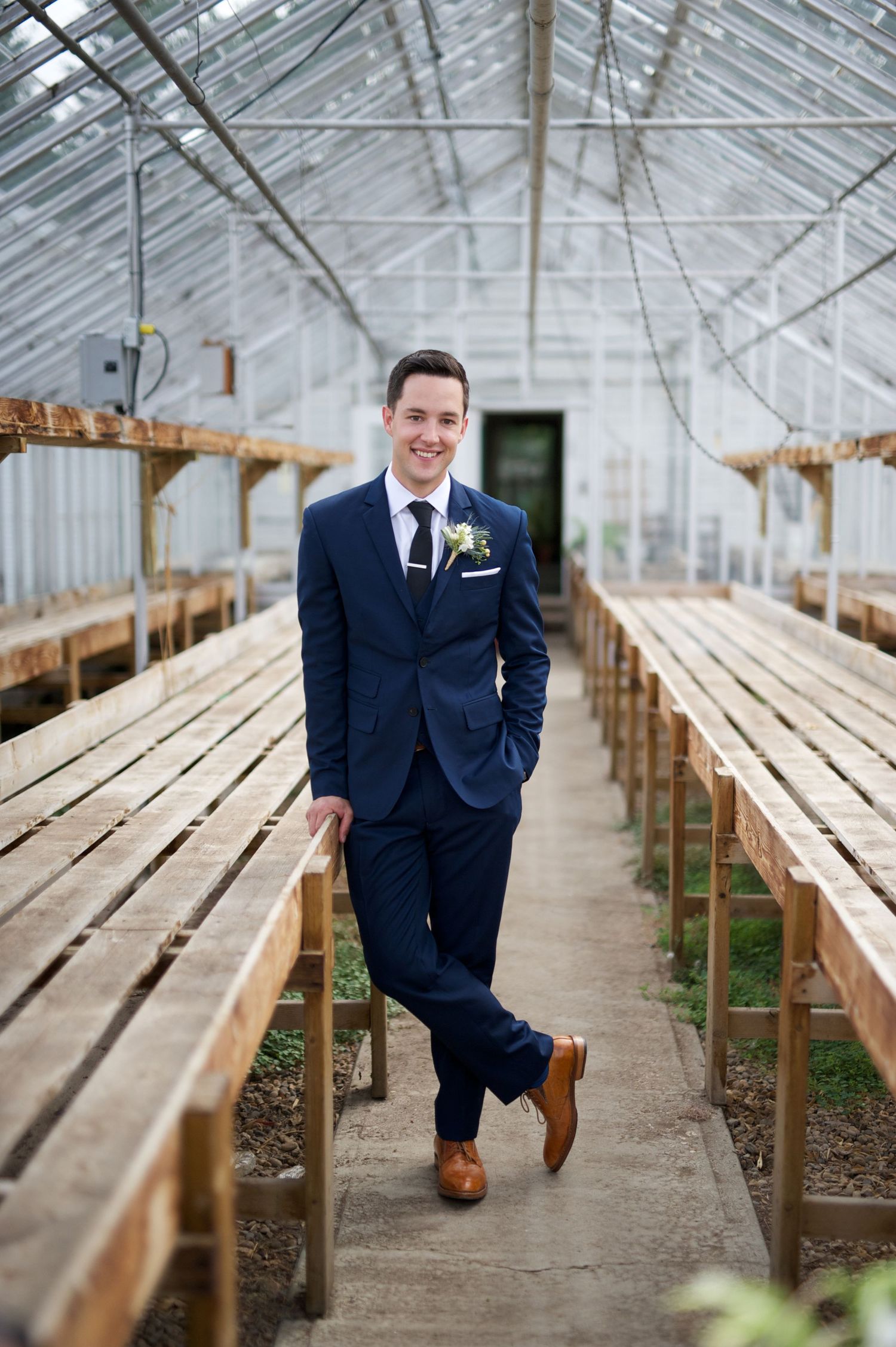 Groom in a navy suit leans confidently against a bench row inside a glass greenhouse.