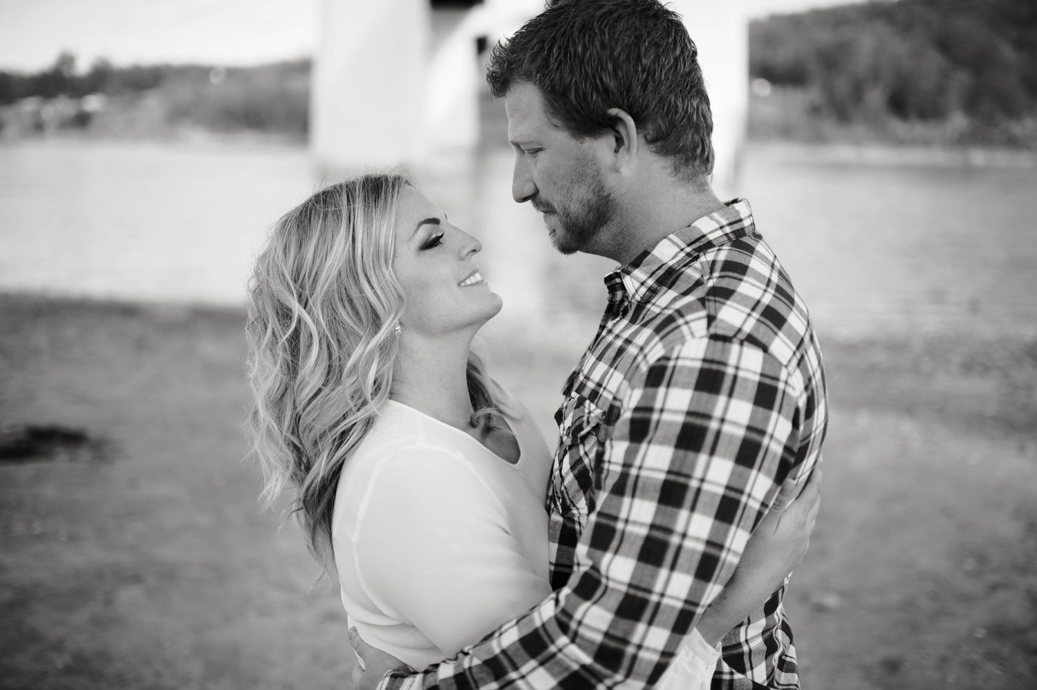 Black and white engagement portrait of a couple standing close on a sandy riverside, sharing a soft gaze under a bridge.