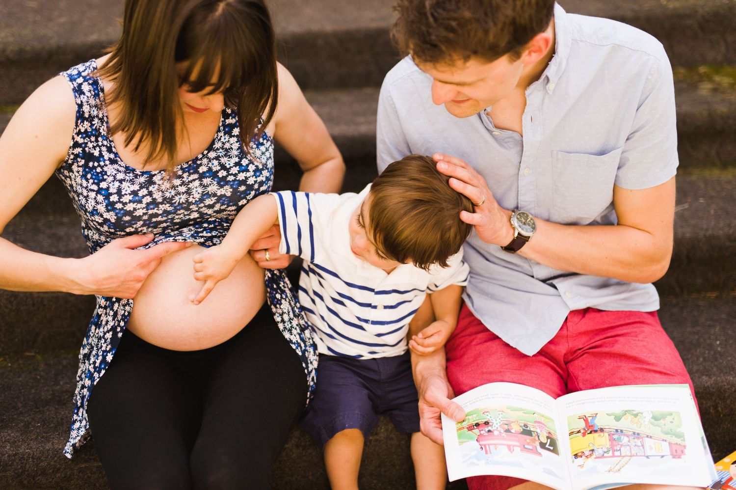Family sitting on front steps reading a book, toddler reaching out to touch mom’s pregnant belly.