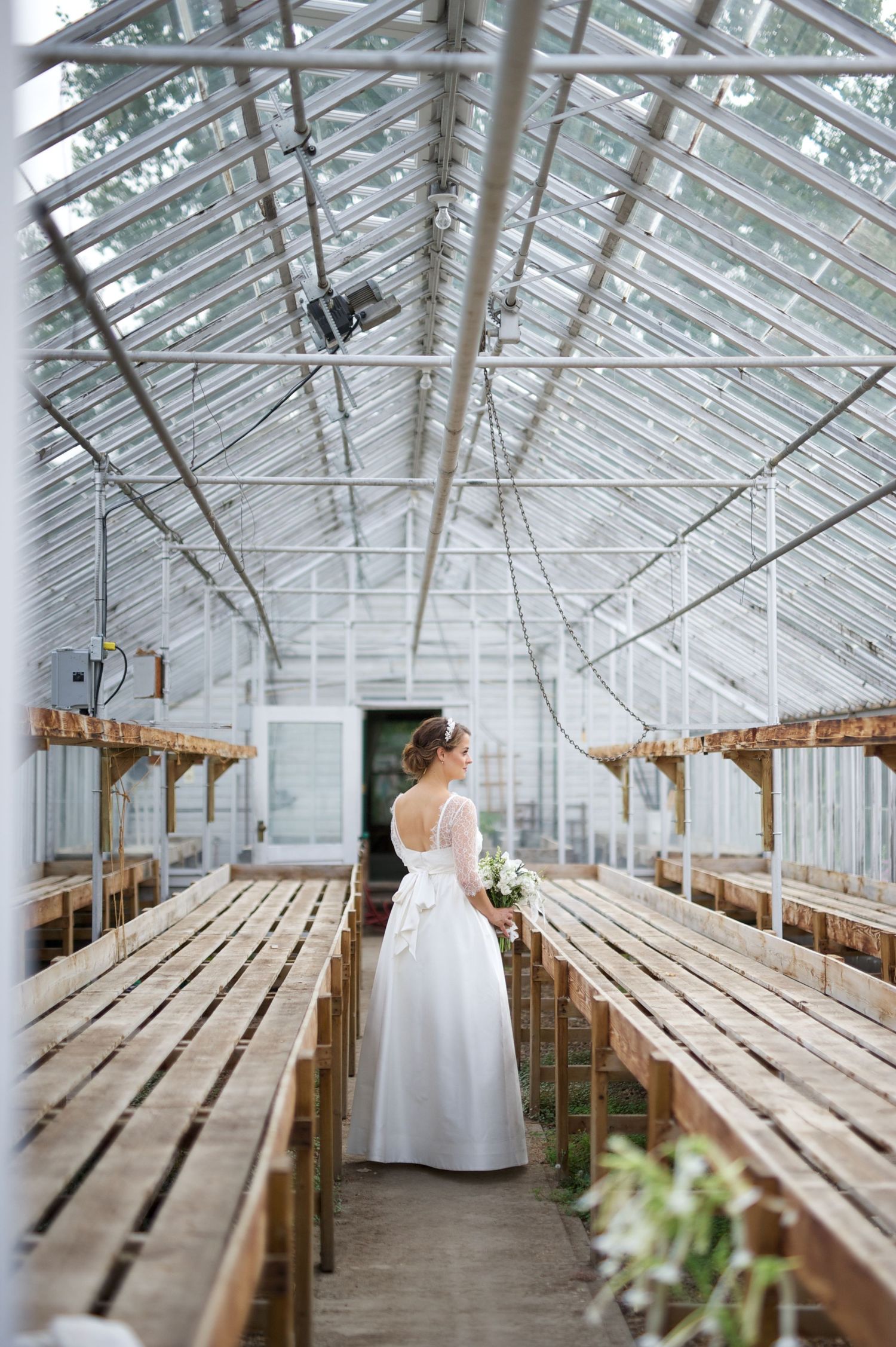 Bride in a flowing gown stands alone in the greenhouse aisle, looking upward and holding her bouquet.