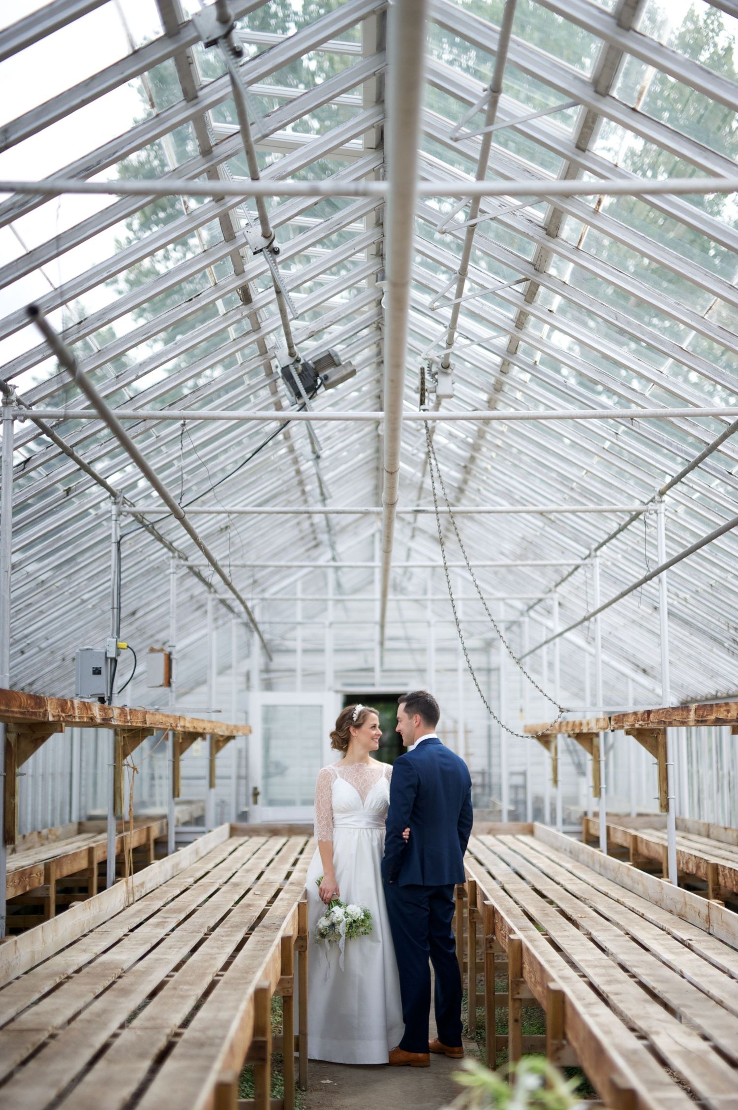 A quiet kiss between newlyweds in a greenhouse aisle. Still, romantic, and softly lit with rustic charm.