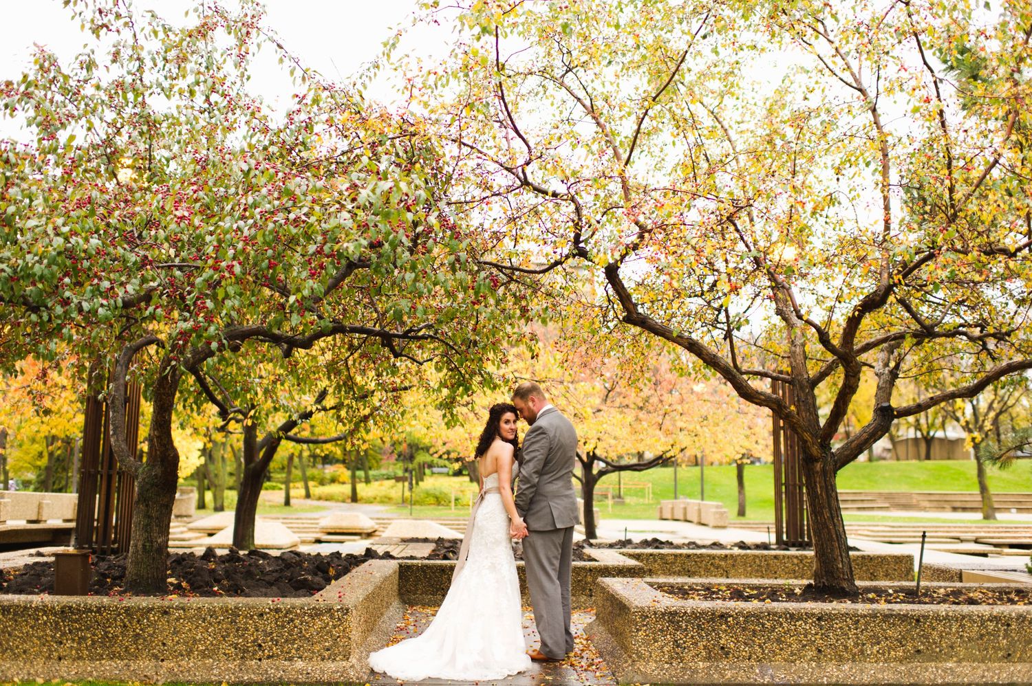 Wedding couple kissing in a park lined with colorful autumn trees, standing atop a stone walkway with soft golden light all around.
