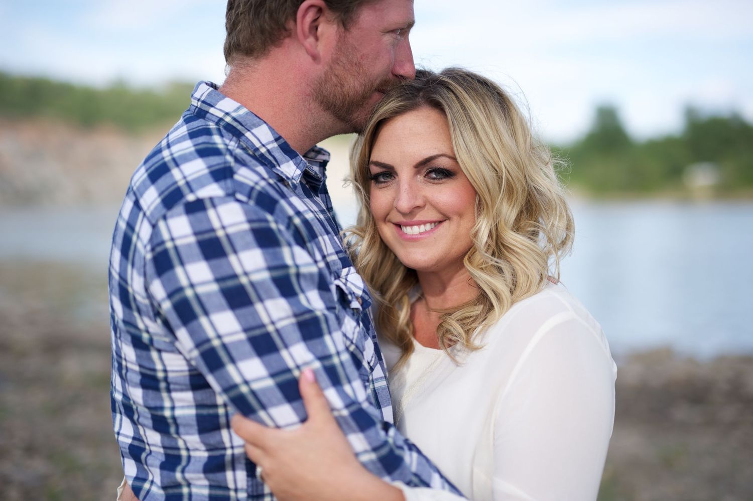 Engagement photo of a couple standing by the river, woman smiling at the camera while embraced by her partner in a plaid shirt.