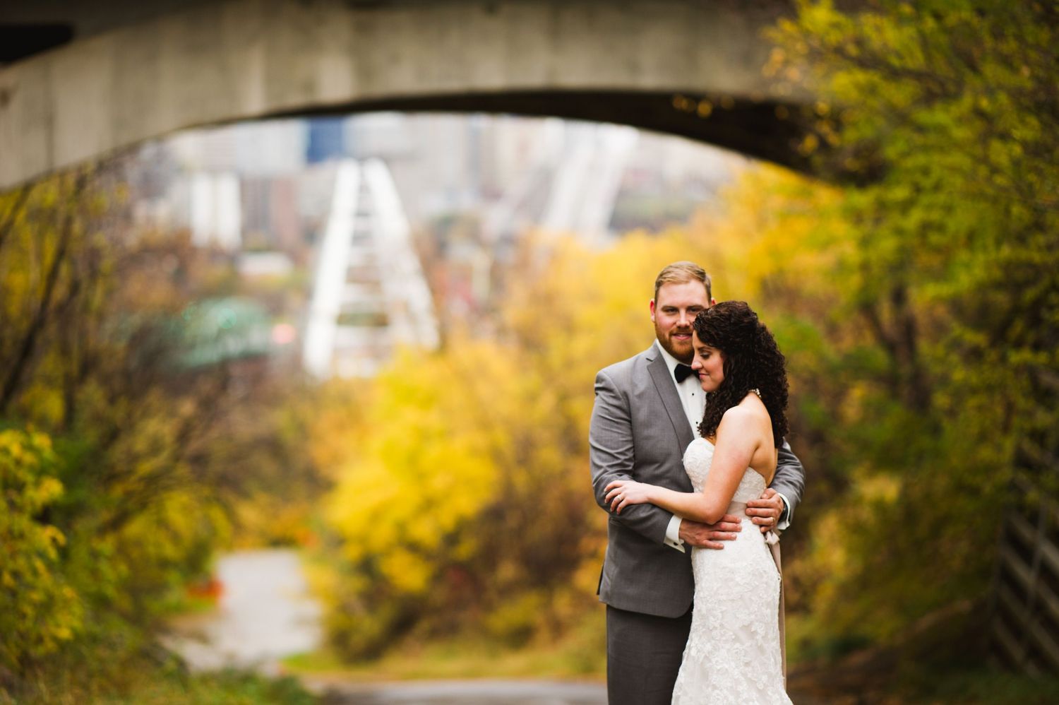 Bride and groom embracing in front of a vibrant autumn backdrop, with the skyline and bridge visible through the trees beneath