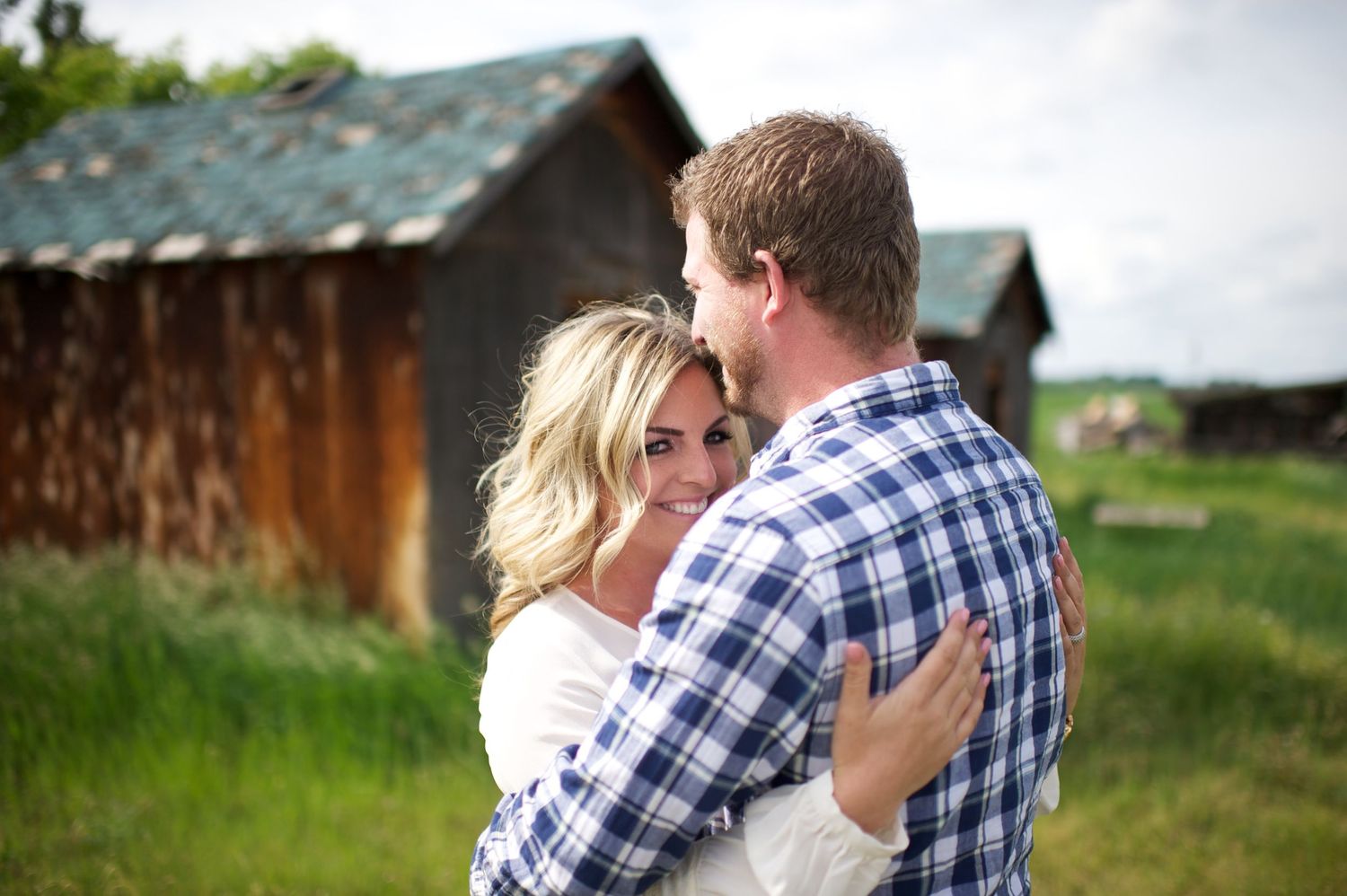 Woman looking over her partner’s shoulder as they embrace in front of rusted barn walls and a grassy field. Rustic Alberta engagement photo.